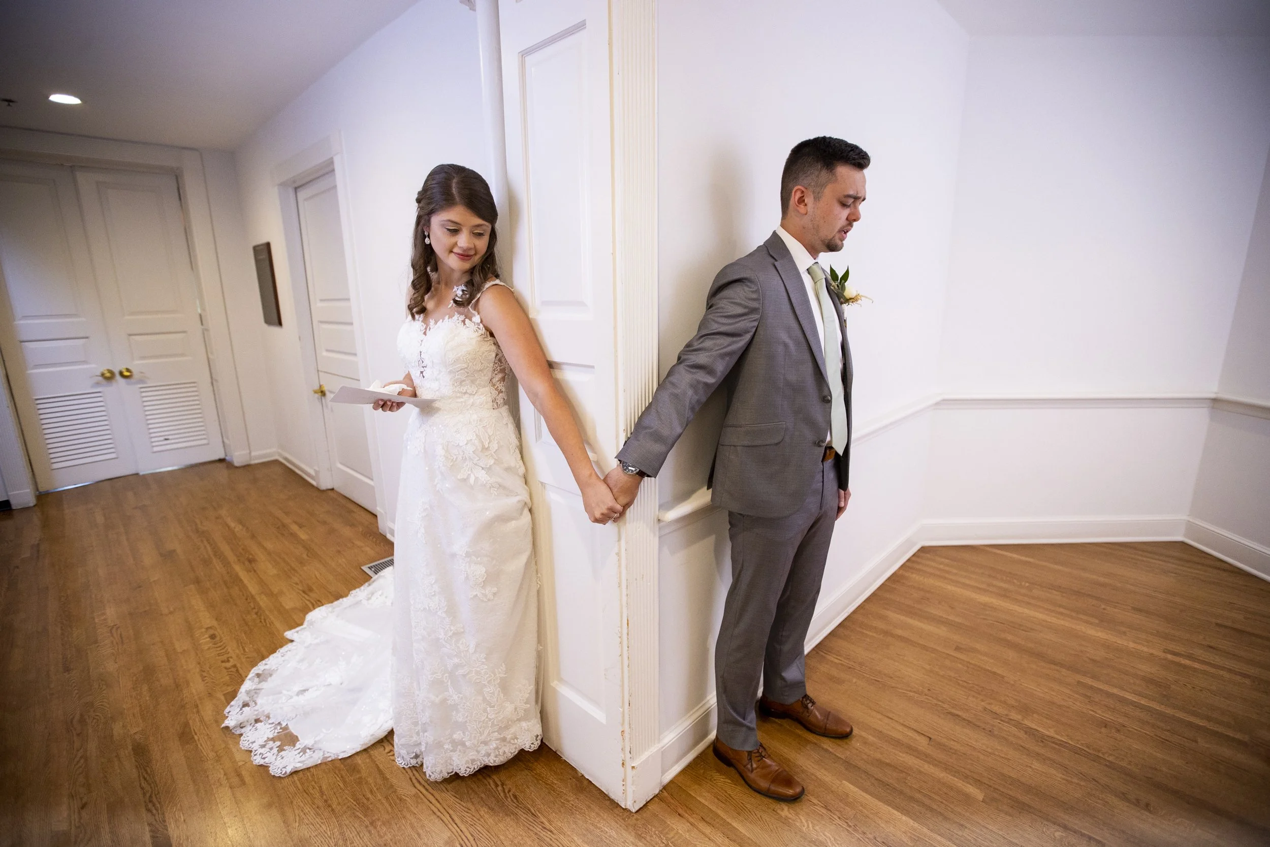 A bride and groom stand back to back, holding hands behind a partition wall in a room with hardwood floors and white walls, during their wedding ceremony.