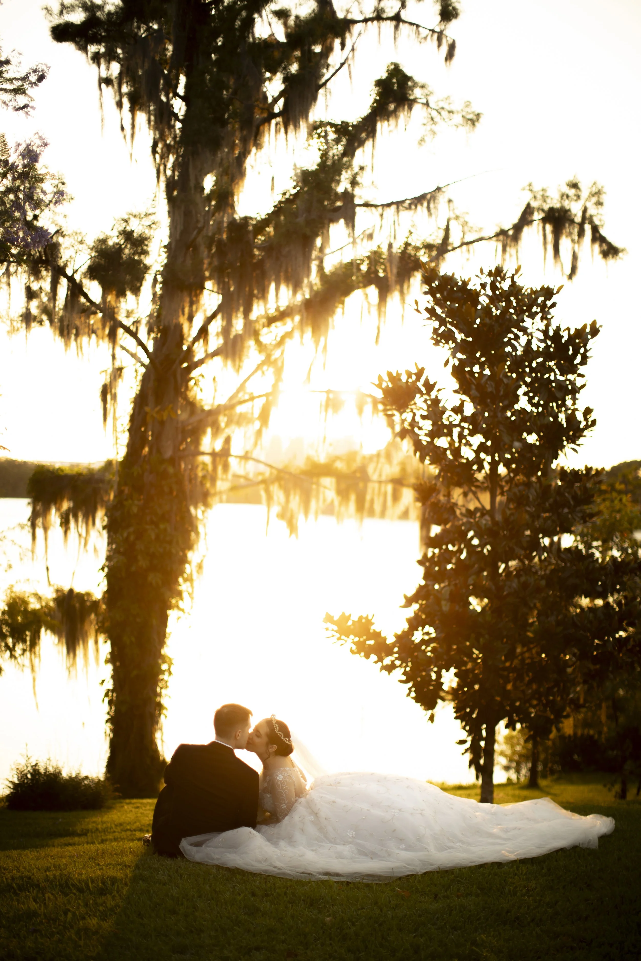 A bride and groom sitting on grass by a lake at sunset, sharing a kiss, surrounded by trees.
