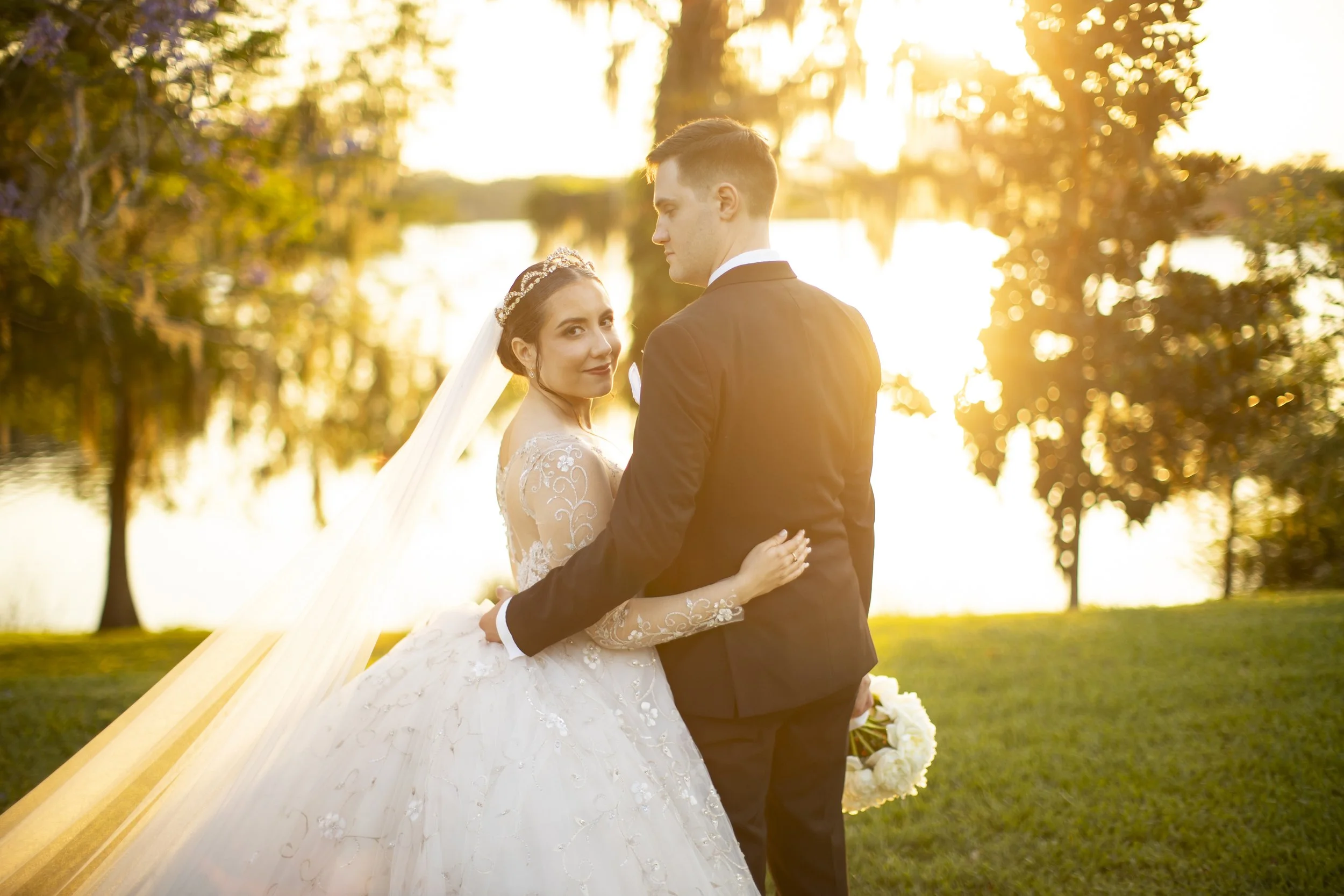 A bride and groom standing outdoors during sunset. The bride is smiling and looking at the camera, wearing a white wedding dress with lace details and a veil, holding a bouquet of white flowers. The groom is facing away, holding the bride around the 