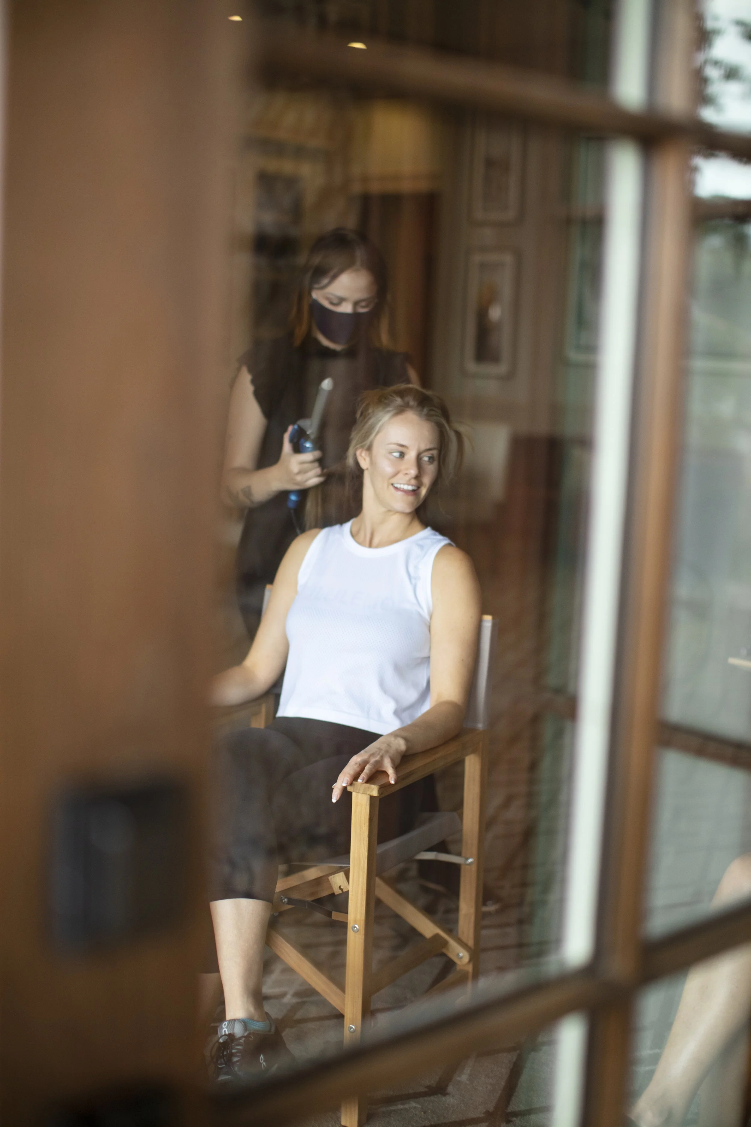 Woman sitting on a wooden chair getting her hair styled by a hairstylist using a curling iron, viewed through a glass window.