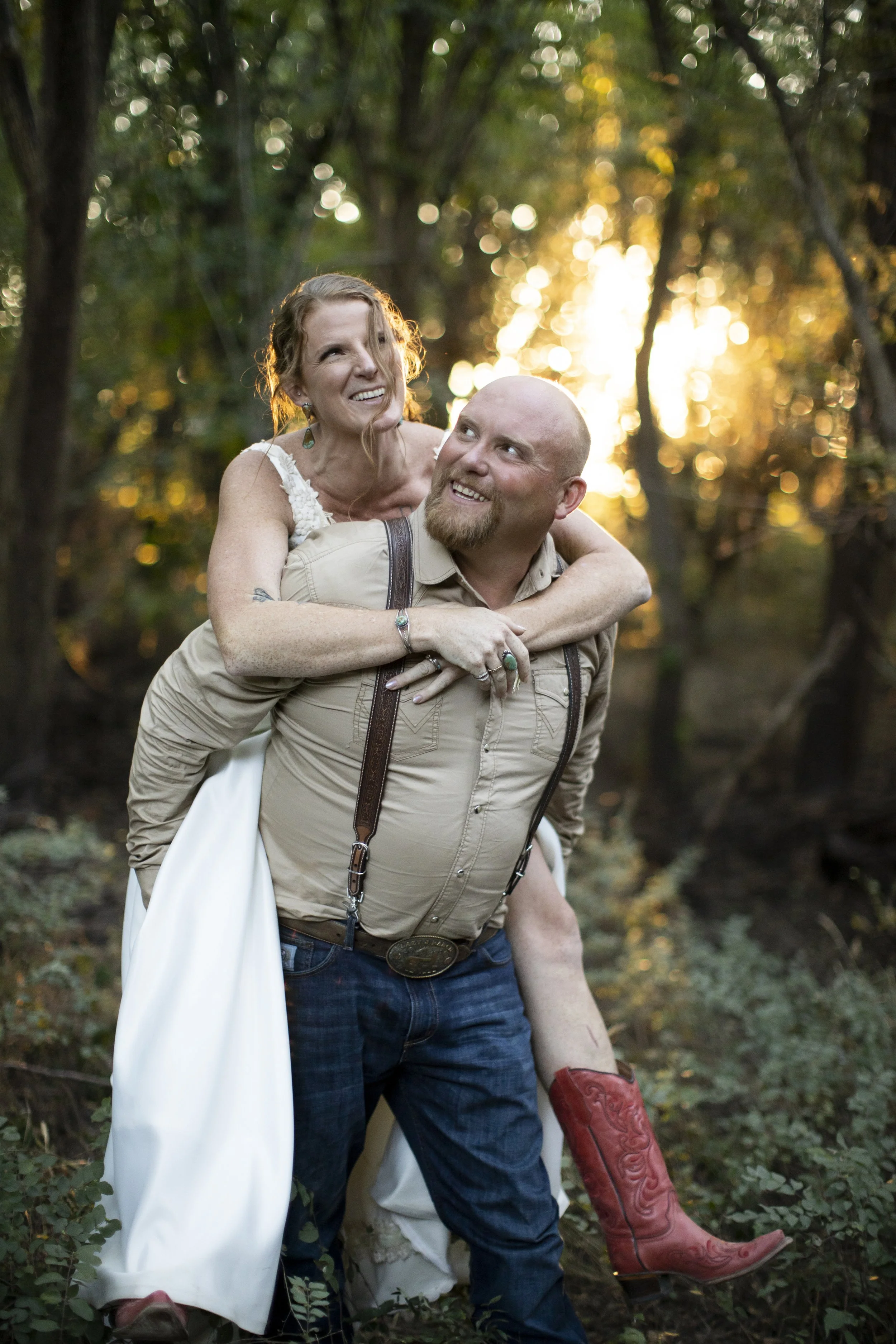 A couple outdoors in a wooded area at sunset, with the woman piggybacking on the man, both smiling and looking at each other. The woman is wearing a white dress, and the man is dressed in a beige shirt with suspenders. The scene has a warm, golden gl