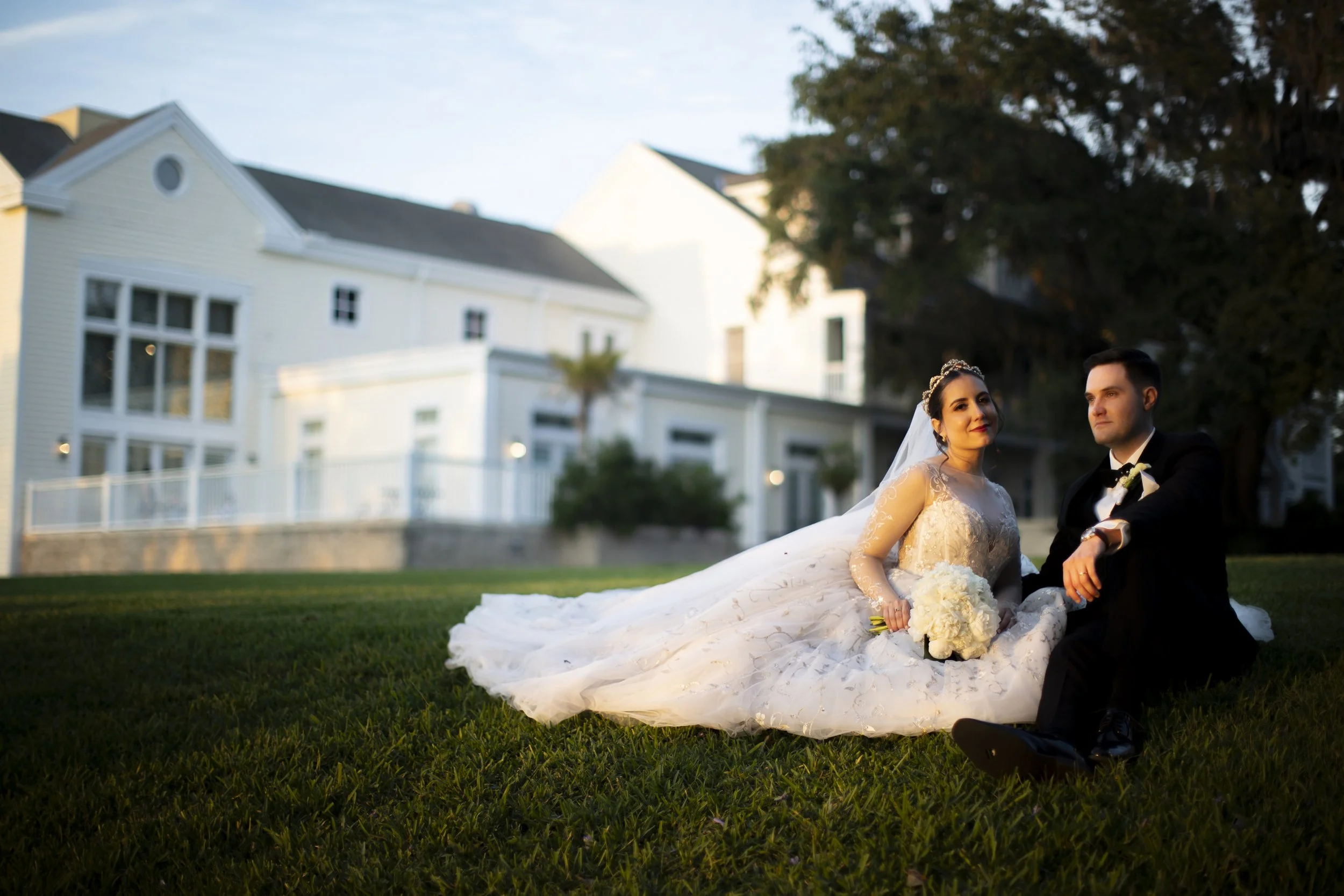 A bride and groom sitting on the grass outside, with a modern white house and trees in the background during sunset.