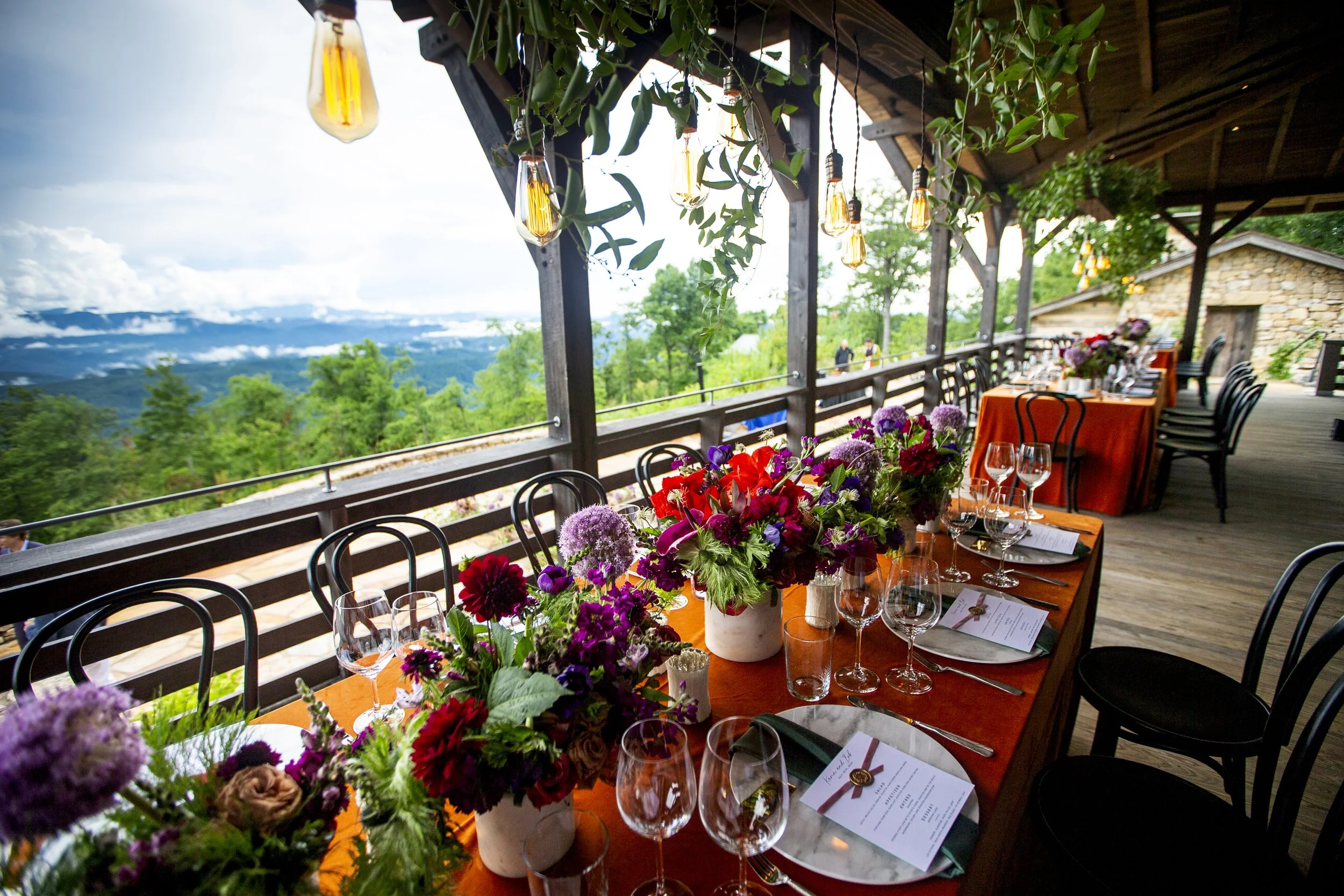 Elegant outdoor dining table set with colorful floral arrangements, wine glasses, and menus under string lights on a covered balcony with a scenic mountain view