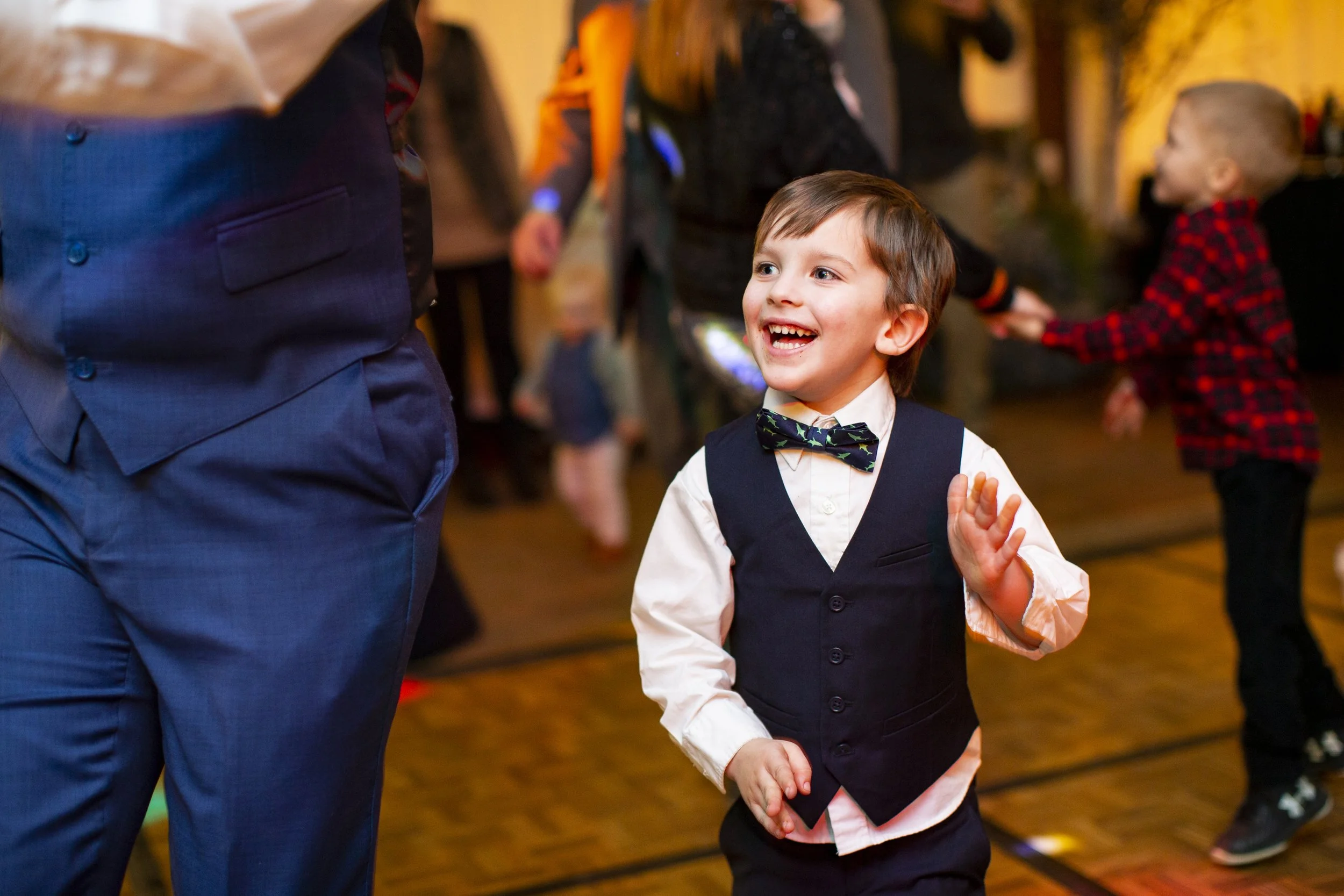 A young boy in formal attire, including a white shirt, black vest, and bow tie, smiling and waving while dancing or playing at a social event, with other children and adults in the background.