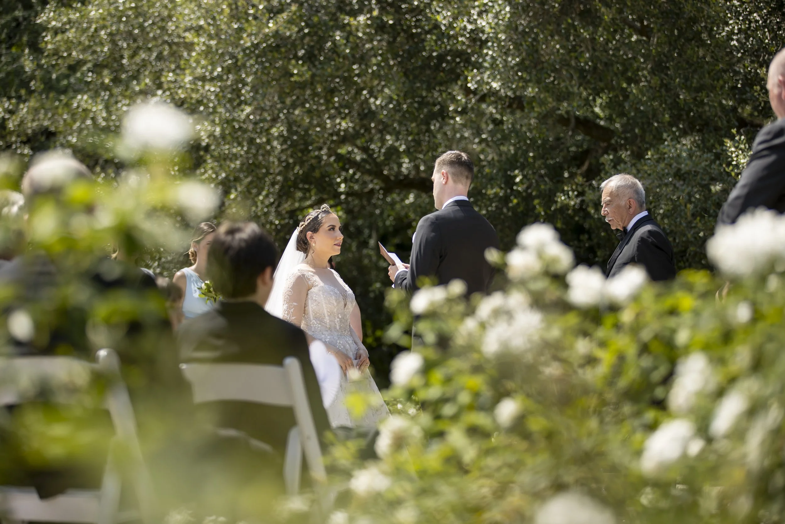 A bride and groom exchange vows at an outdoor wedding ceremony, surrounded by guests and lush greenery.