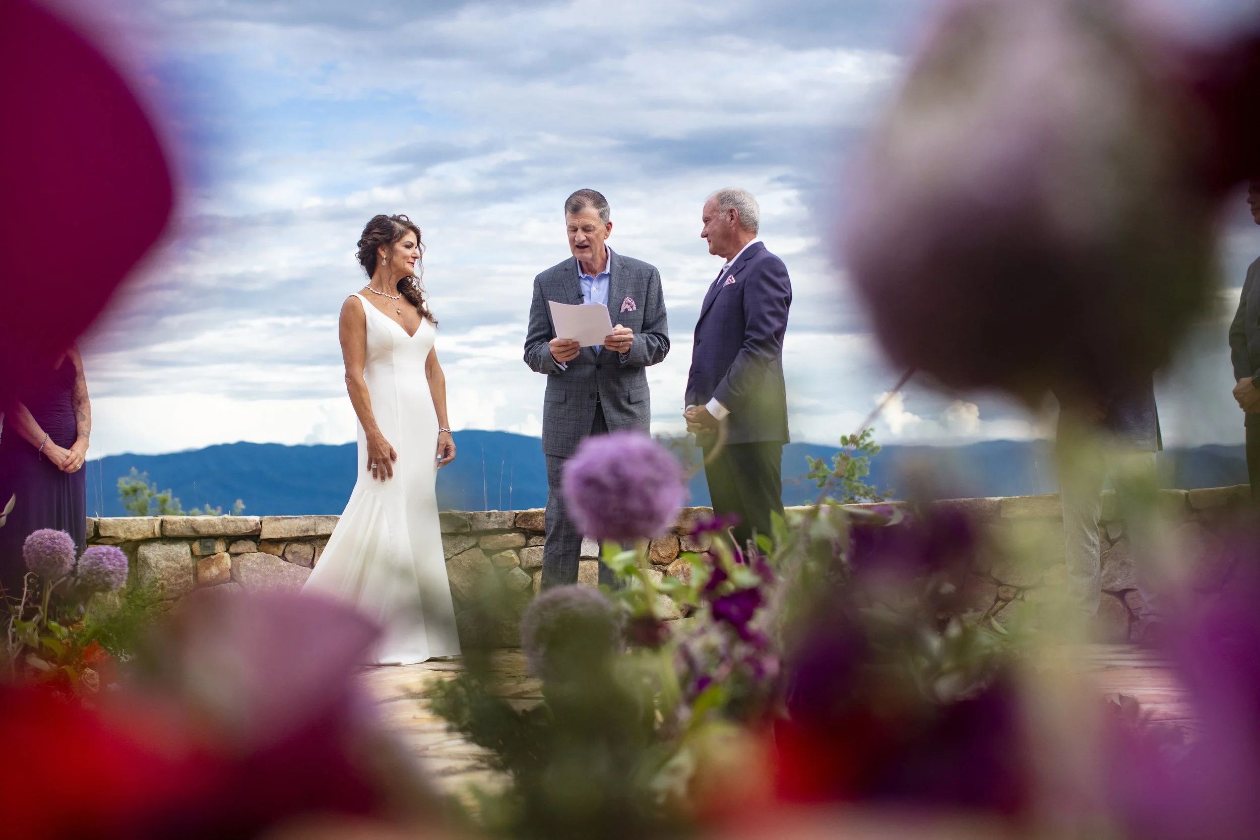 Wedding ceremony outdoors with a bride in a white gown and two men in suits, one holding a paper, with mountains in the background, framed by flowers in the foreground.