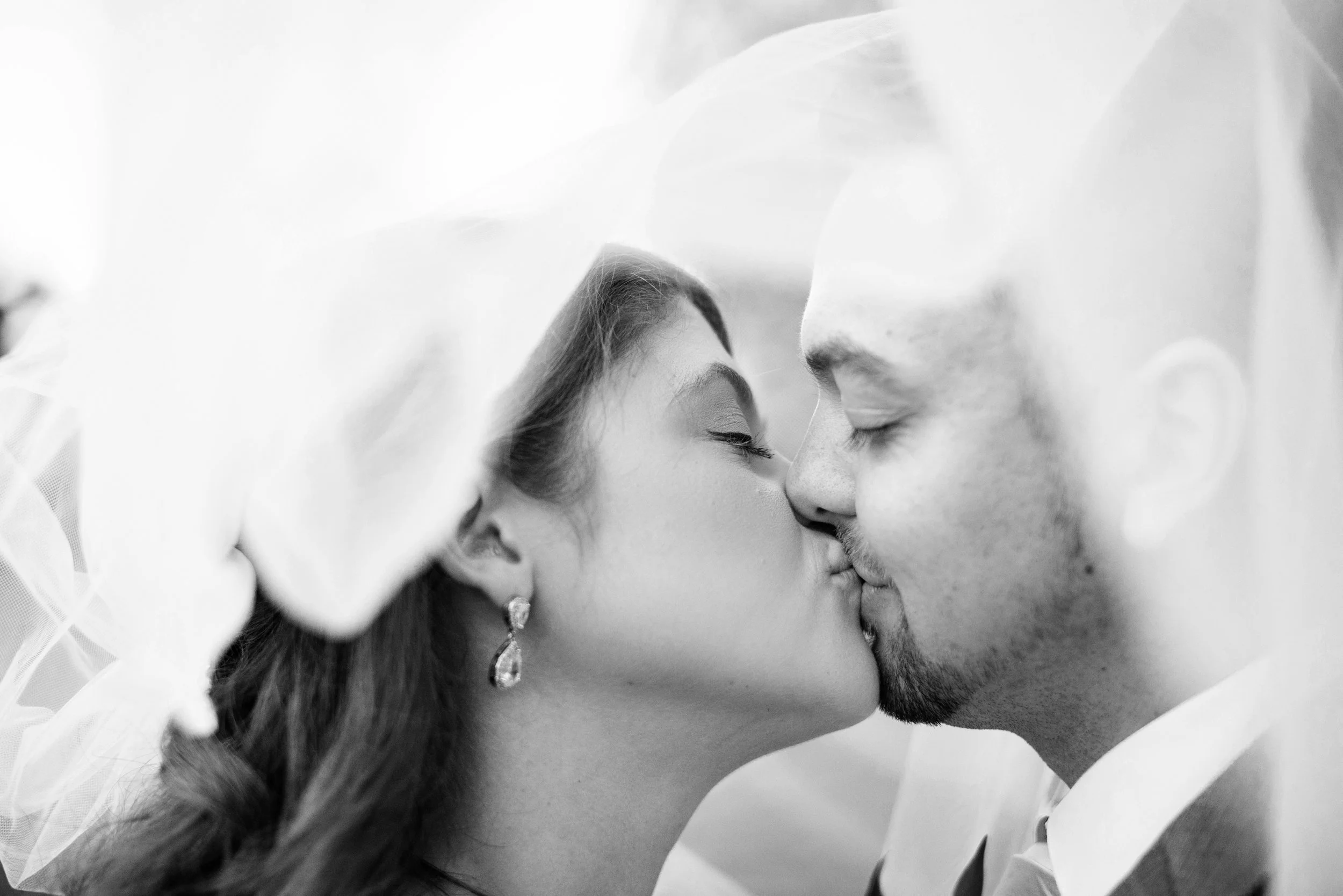 Black-and-white photo of a bride and groom sharing a kiss, with a wedding veil partially covering their faces.