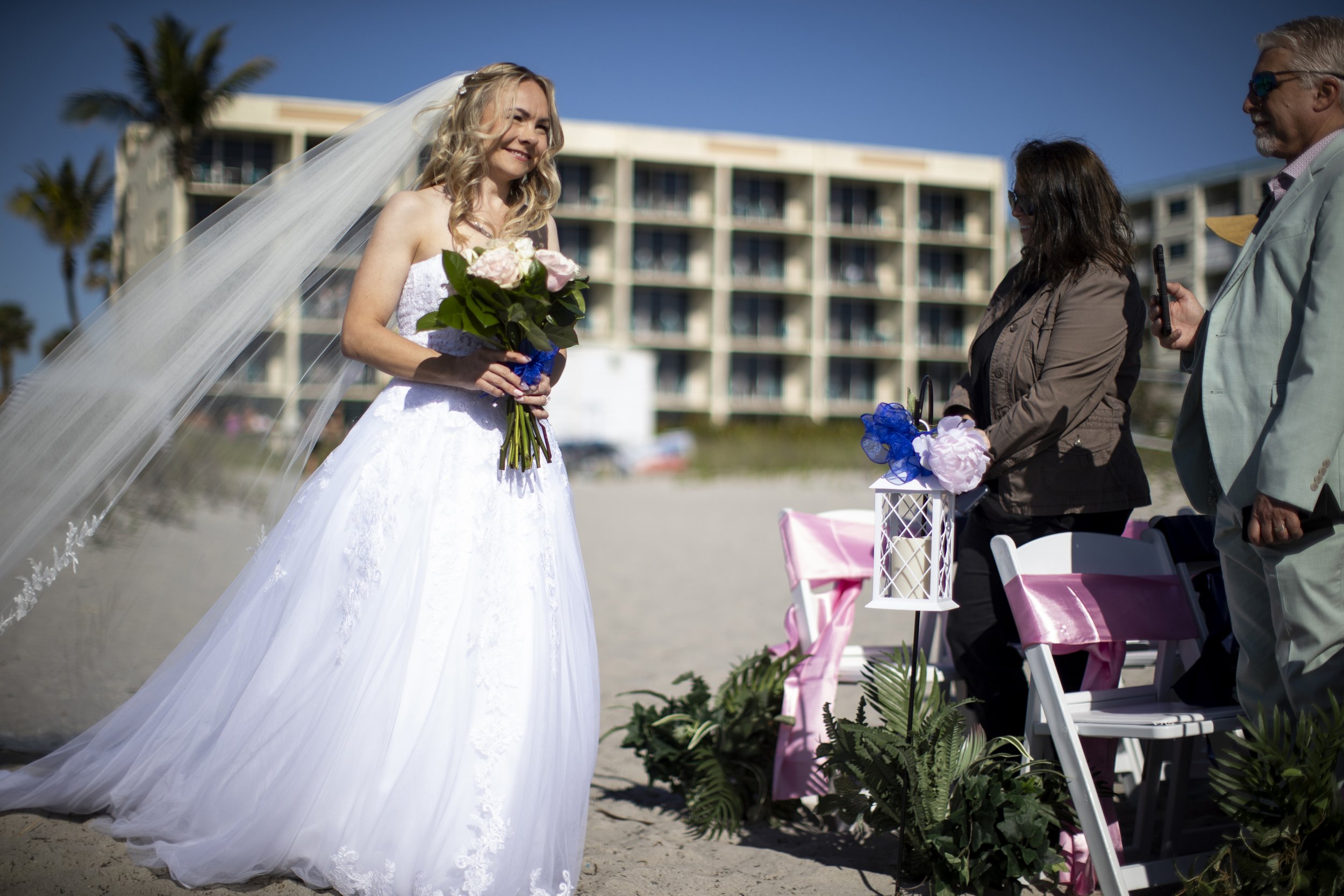 A bride in a white wedding dress holding a bouquet of pink and white flowers on a beach, with a woman and man nearby, and a building and palm trees in the background.