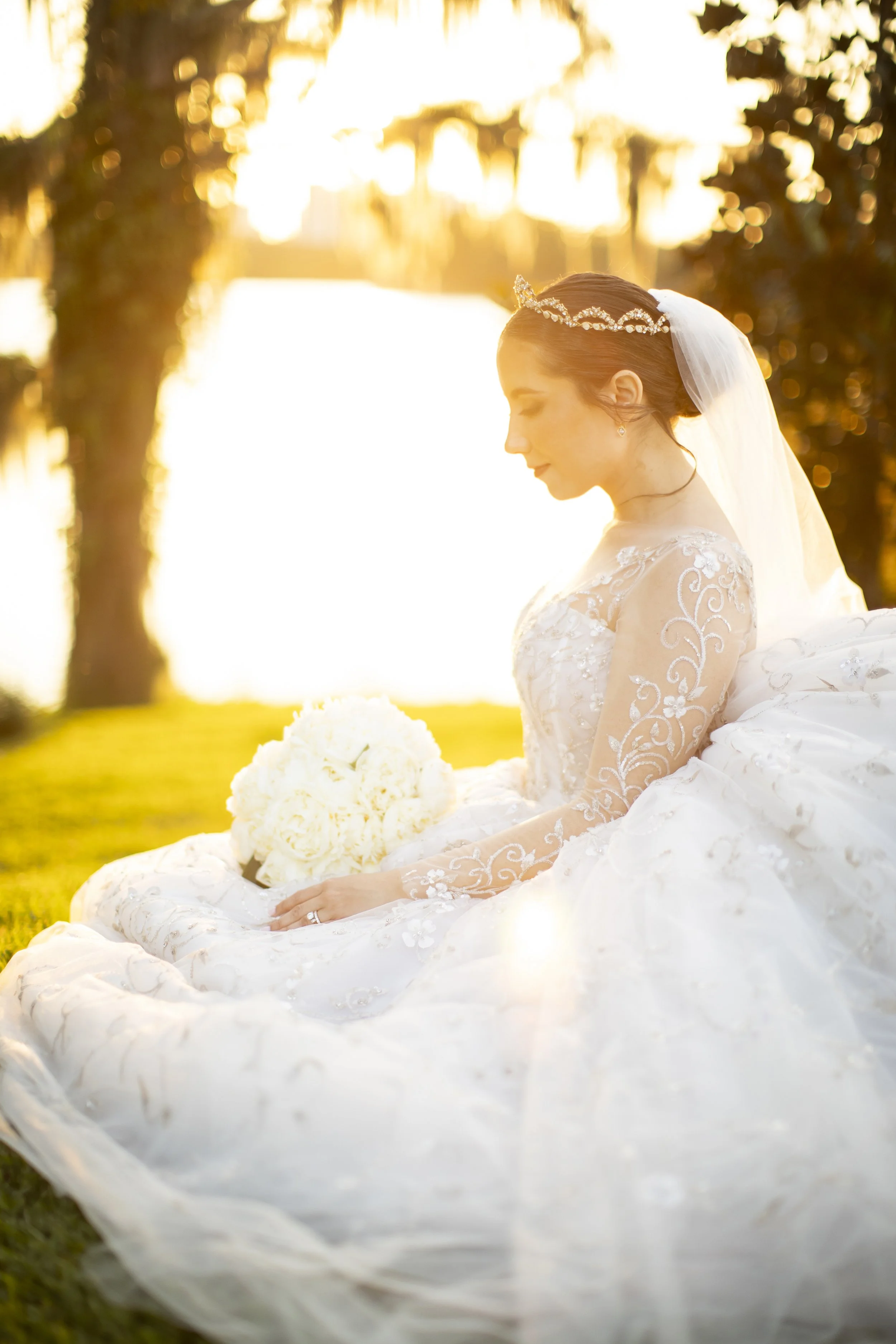 A bride in a white wedding dress with lace details, holding a bouquet of white flowers, sitting on grass outdoors during sunset.