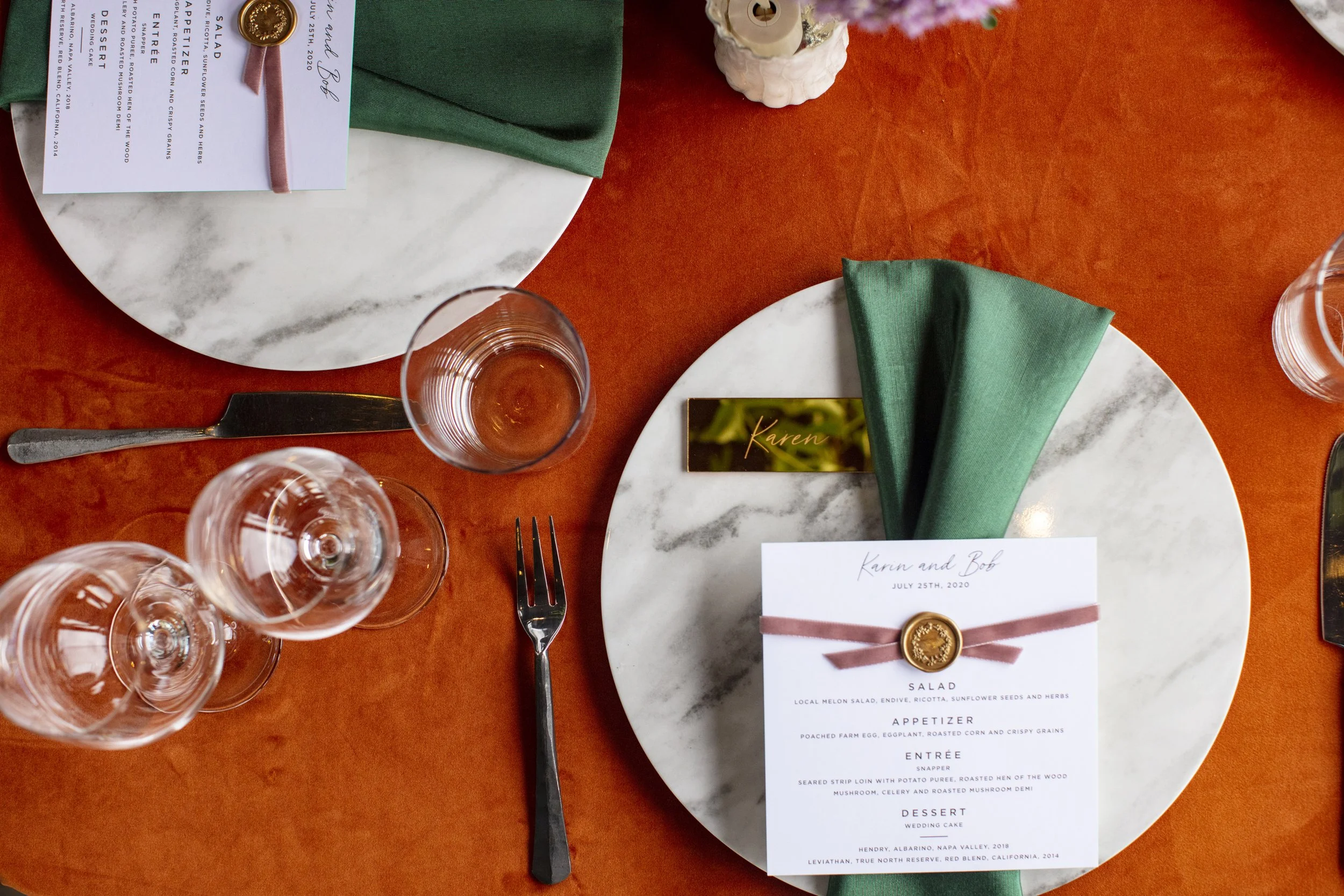 Elegant table setting for a wedding or formal event with a white marble plate, a green cloth napkin, a black place card labeled 'Karen', a menu with a pink ribbon and wax seal, three wine glasses, and cutlery on an orange tablecloth.