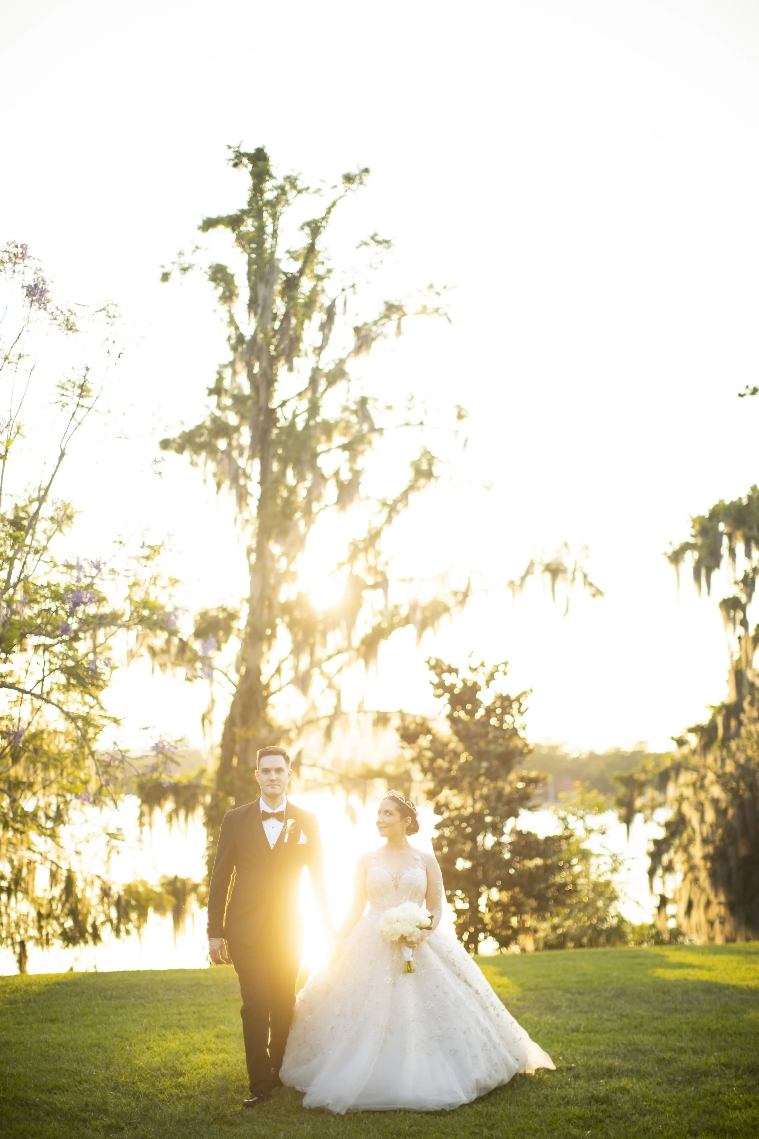 A bride and groom walking hand in hand on a grassy area during sunset, with tall trees and Spanish moss in the background.