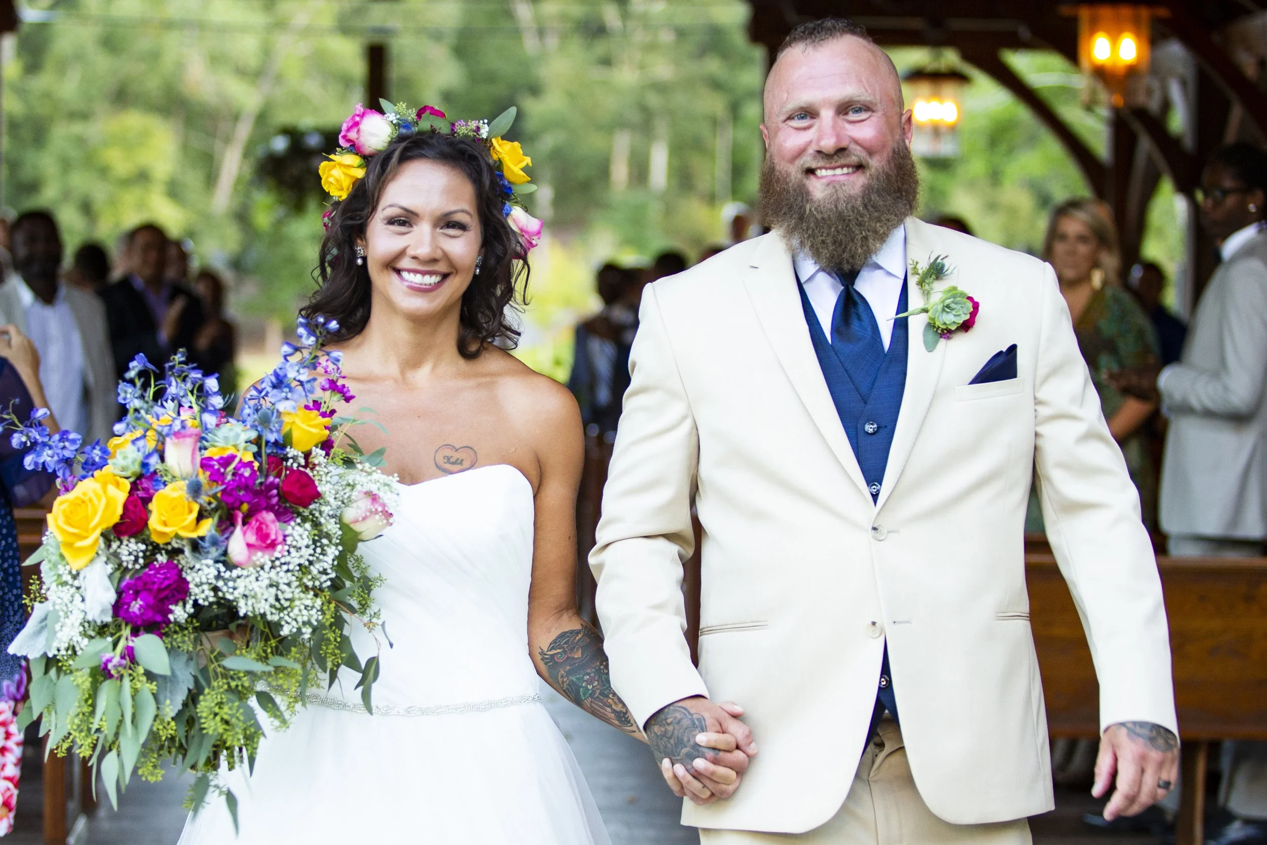 Happy couple at a wedding, holding hands and smiling, with guests in the background.