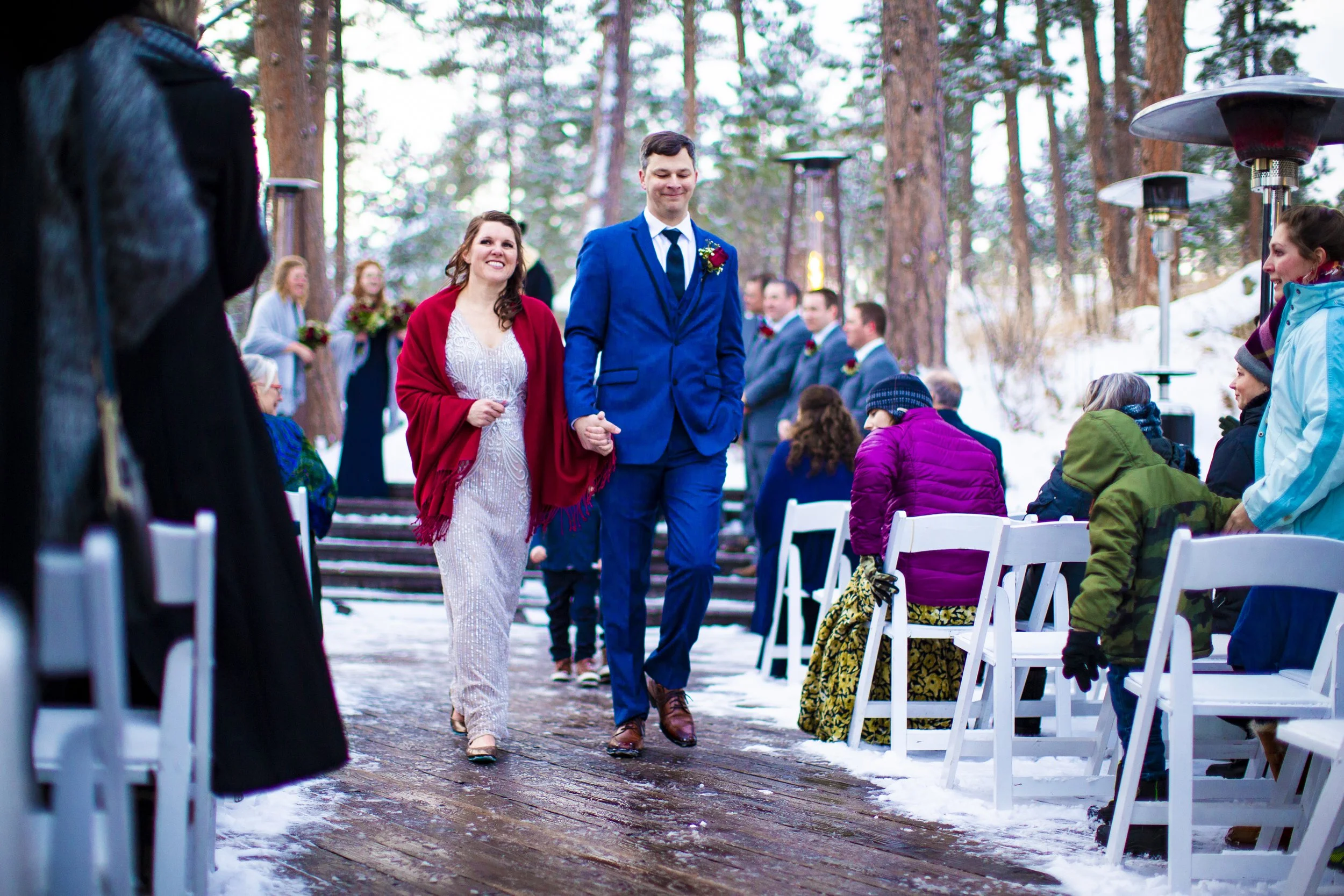 A wedding ceremony taking place outdoors in a snowy forest with pine trees. The bride and groom are walking down the aisle, holding hands. The bride is wearing a white dress and red shawl, and the groom is in a blue suit. Guests are seated on white c