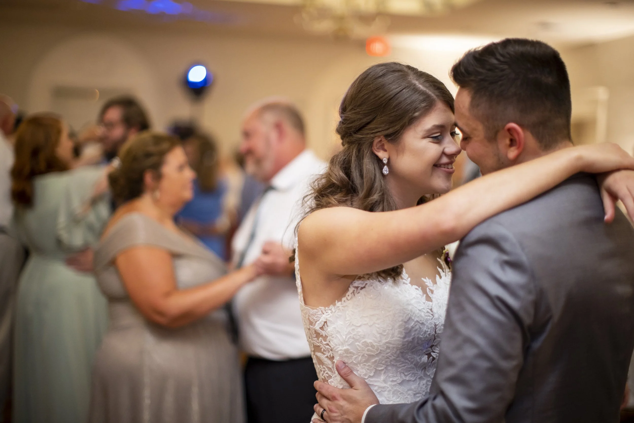 A couple dancing closely at a wedding, smiling and holding each other. The woman is wearing a lace wedding dress, and the man is in a gray suit. Other guests are dancing in the background.