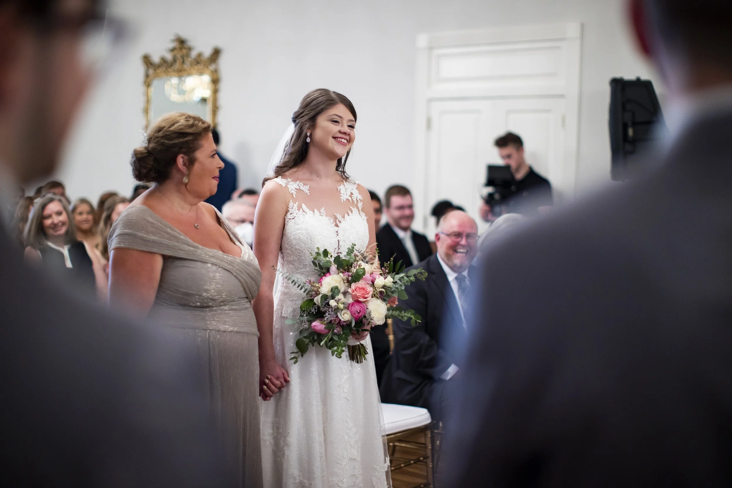 Bride holding a bouquet, standing with a woman at a wedding ceremony surrounded by seated guests.