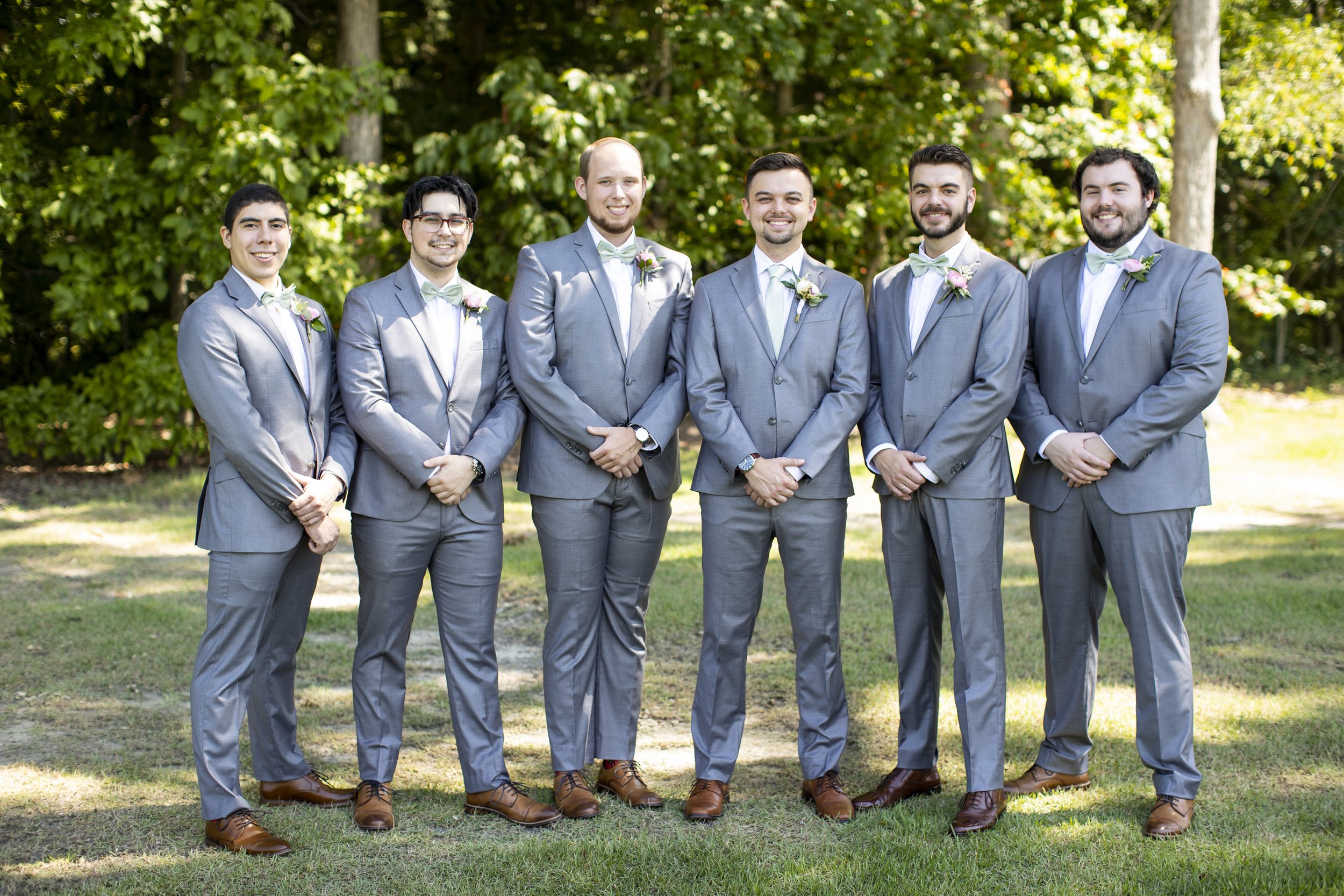 Group of seven men in gray suits with light-colored bow ties and boutonnières, standing outdoors on grass, smiling for a photo with trees in the background.