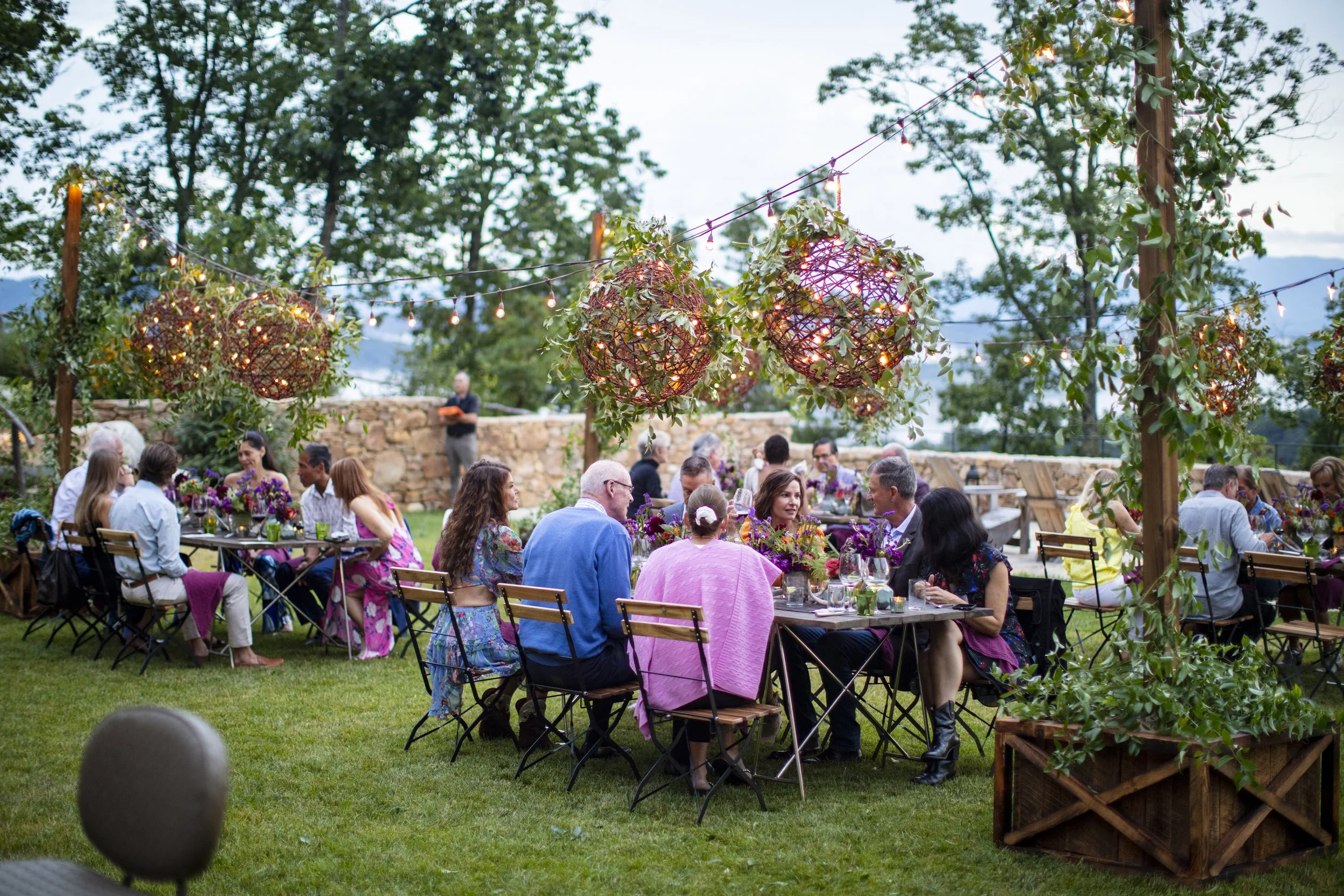 People dining outdoors at a garden party, sitting at long tables decorated with flowers, under string lights and hanging lanterns.