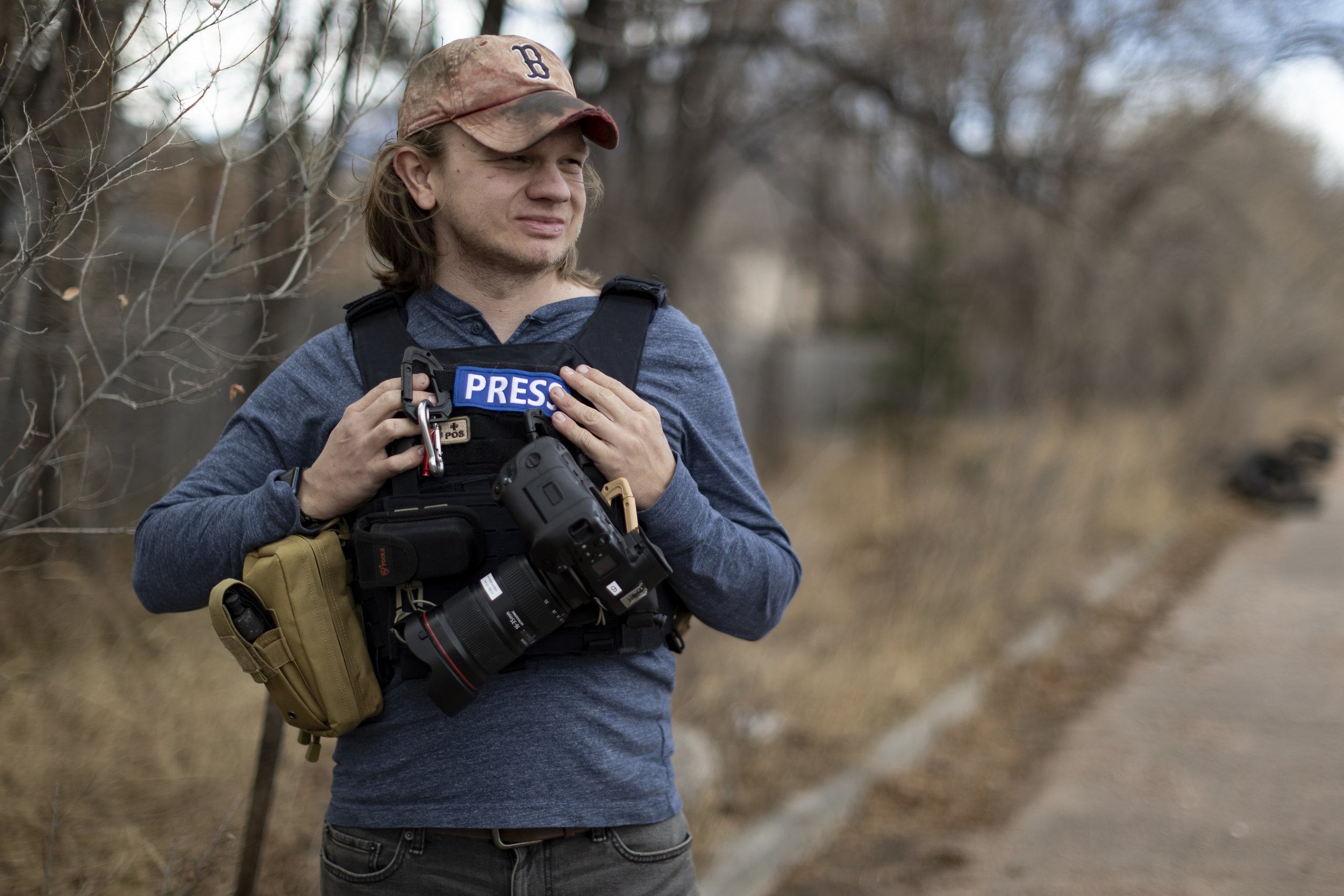 A male journalist standing outdoors near a dirt road, wearing a press vest and holding a camera with a telephoto lens, with a patch labeled 'PRESS' on his vest.