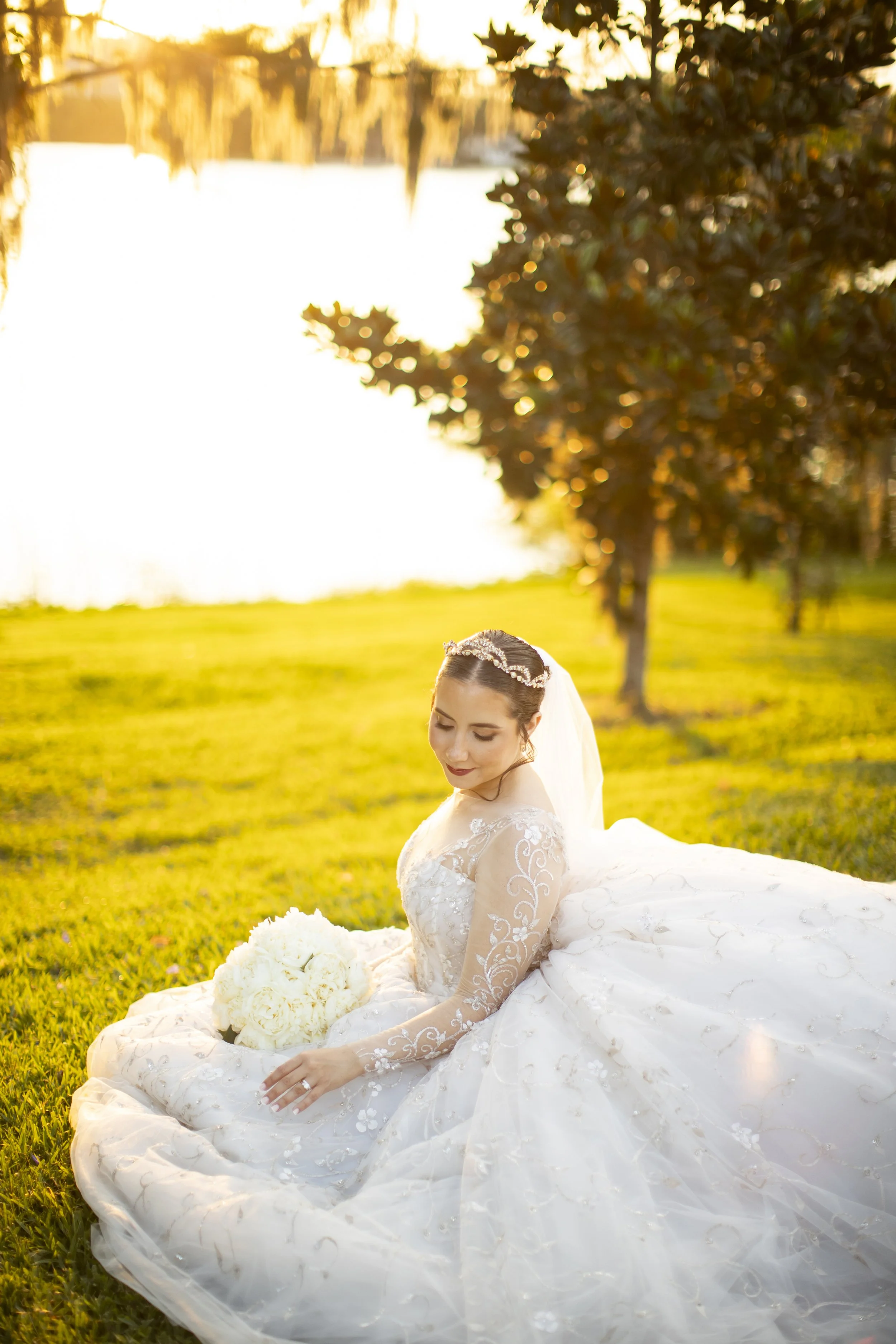 A bride in a white wedding gown sitting on grass near a lake at sunset, holding a bouquet of white flowers.