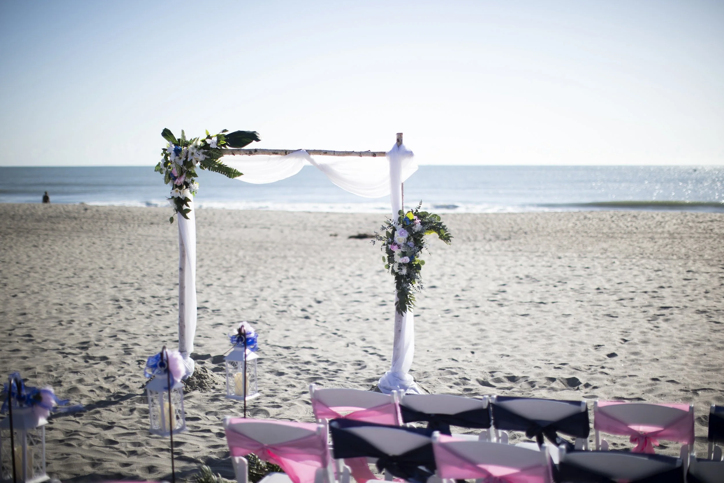 Beach wedding arch decorated with flowers and white fabric, facing the ocean with chairs arranged for guests in front.