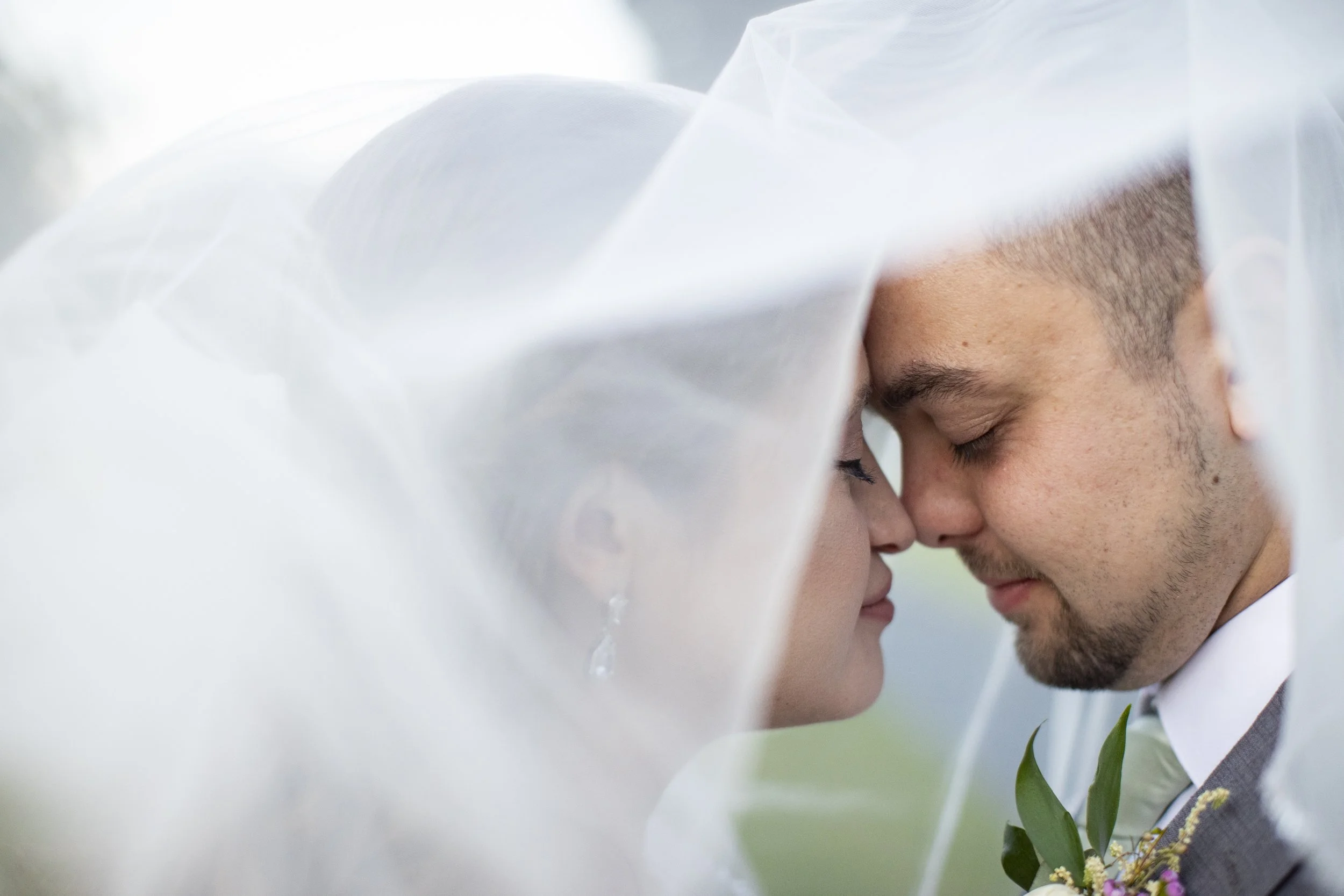 A couple under a wedding veil with their foreheads touching and eyes closed, smiling gently.