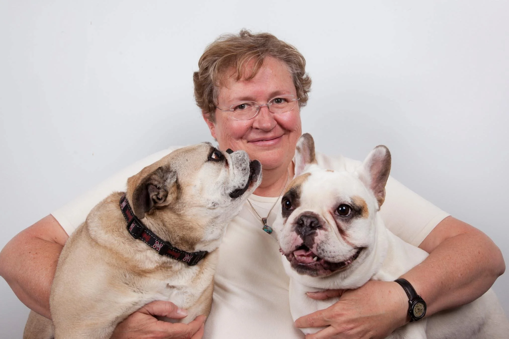 A woman holding two French bulldogs, one on each arm, against a plain white background.