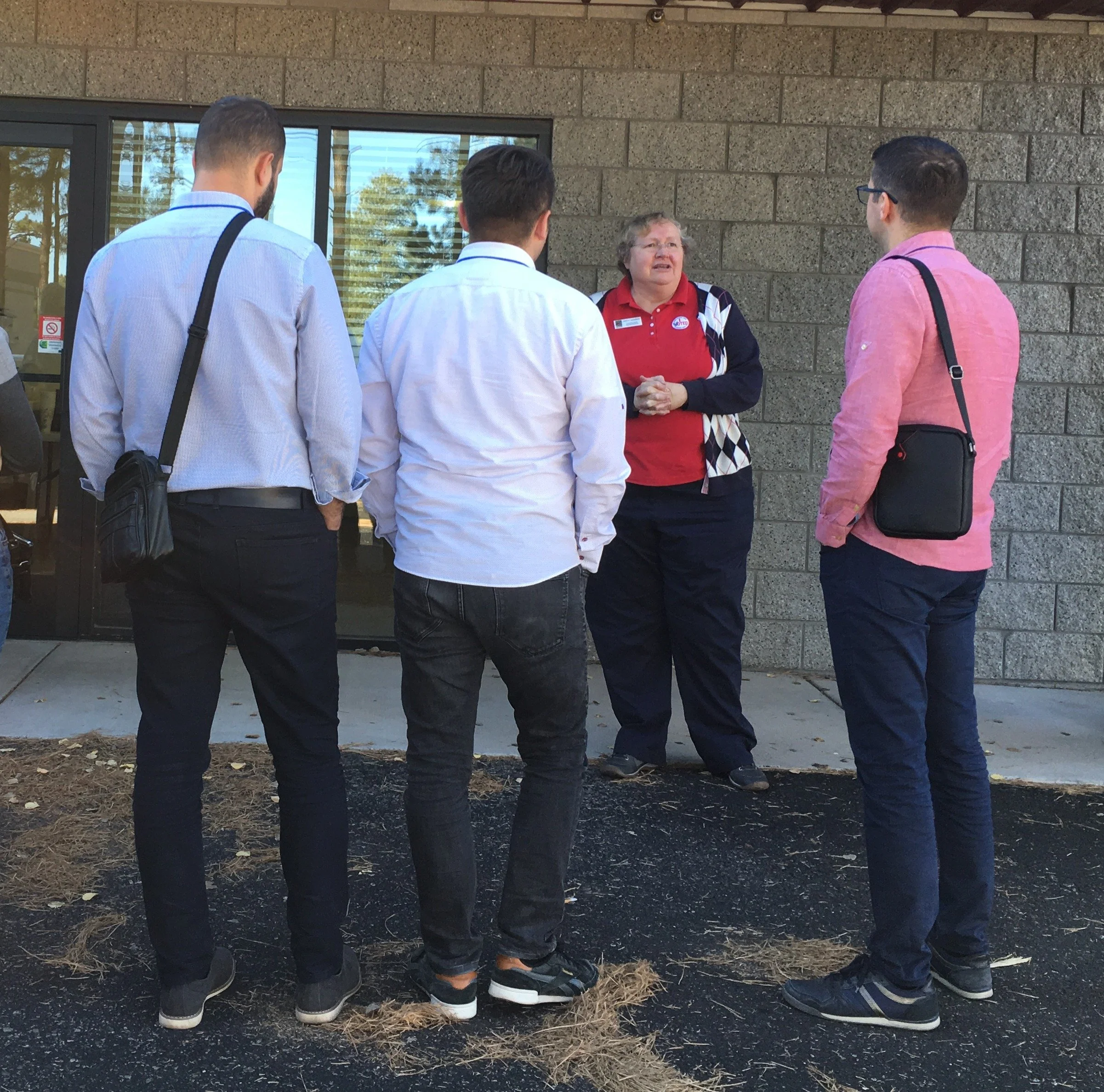 Four people standing outdoors in front of a building, engaged in conversation with a woman wearing a red and black shirt.