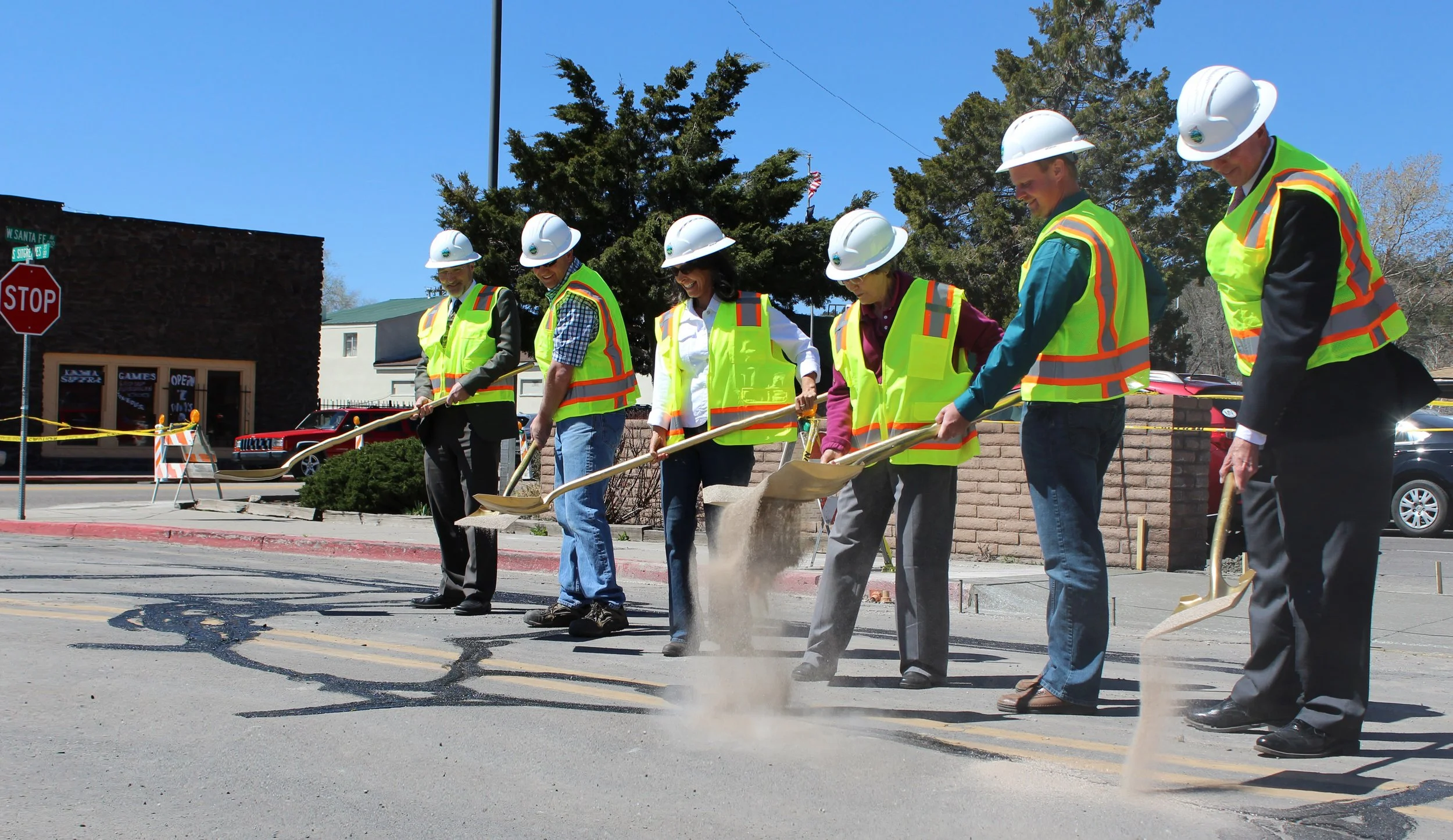 Group of seven construction workers wearing safety vests and hard hats, working together on road repair and filling potholes with sand using shovels.