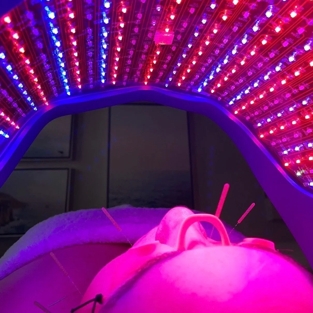 Close-up of a person receiving LED red light therapy on face, under a curved array of colorful LED lights at a clinic or spa setting.