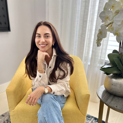 A woman with long brown hair smiling, sitting in a yellow armchair in a well-lit room, with white curtains and a plant with white flowers nearby.