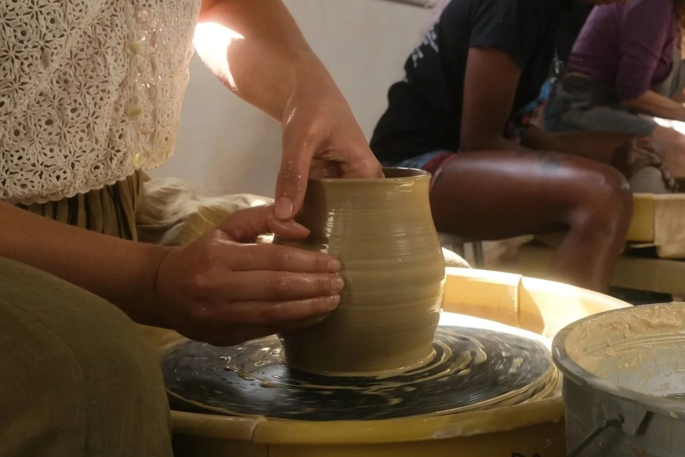 A person shaping a ceramic vase on a pottery wheel with wet hands, surrounded by other people in a pottery class.