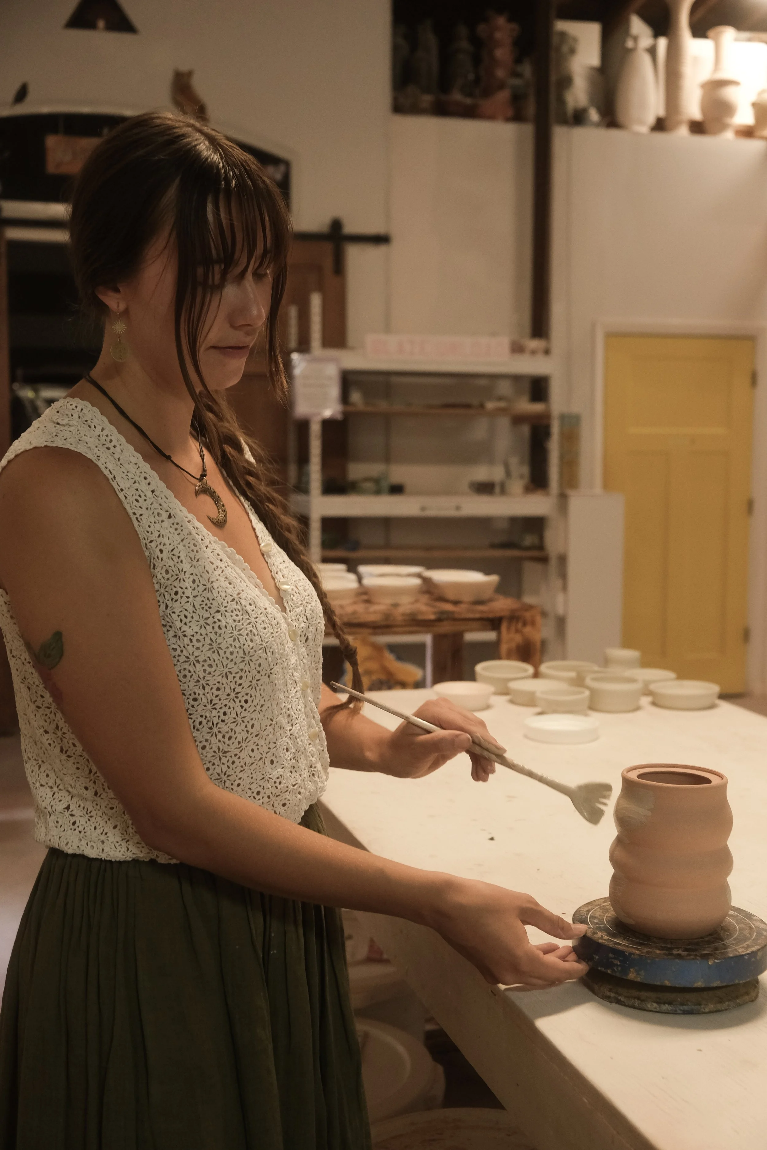A woman with long brown hair, wearing a white crocheted top and a green skirt, is shaping a ceramic pot on a pottery wheel in a pottery studio. She is holding a tool in her right hand and resting her left hand on the pottery. Several other pottery pieces are visible in the background.