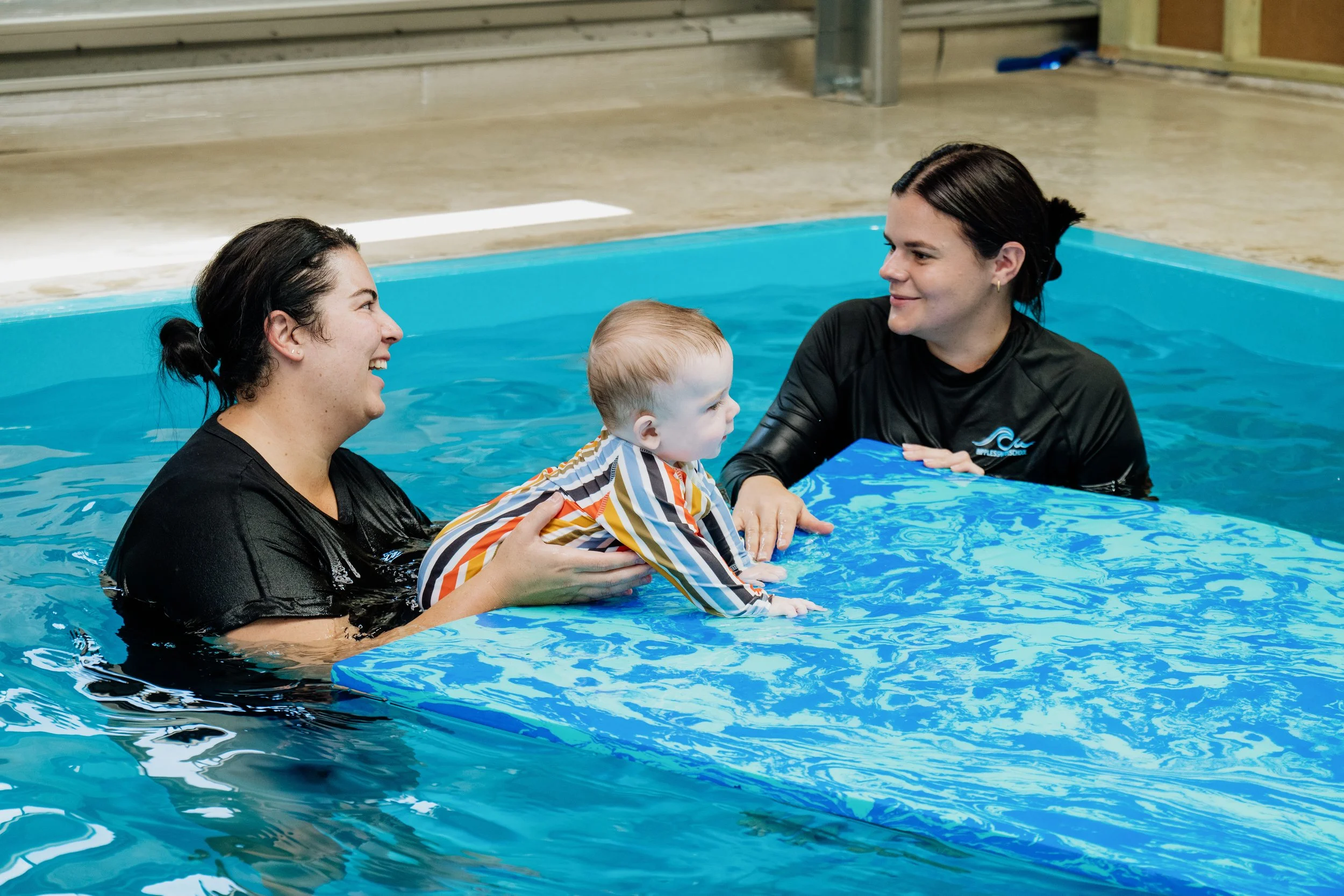 Two women in black shirts assist a young child in a striped shirt during a swimming lesson in an indoor pool.