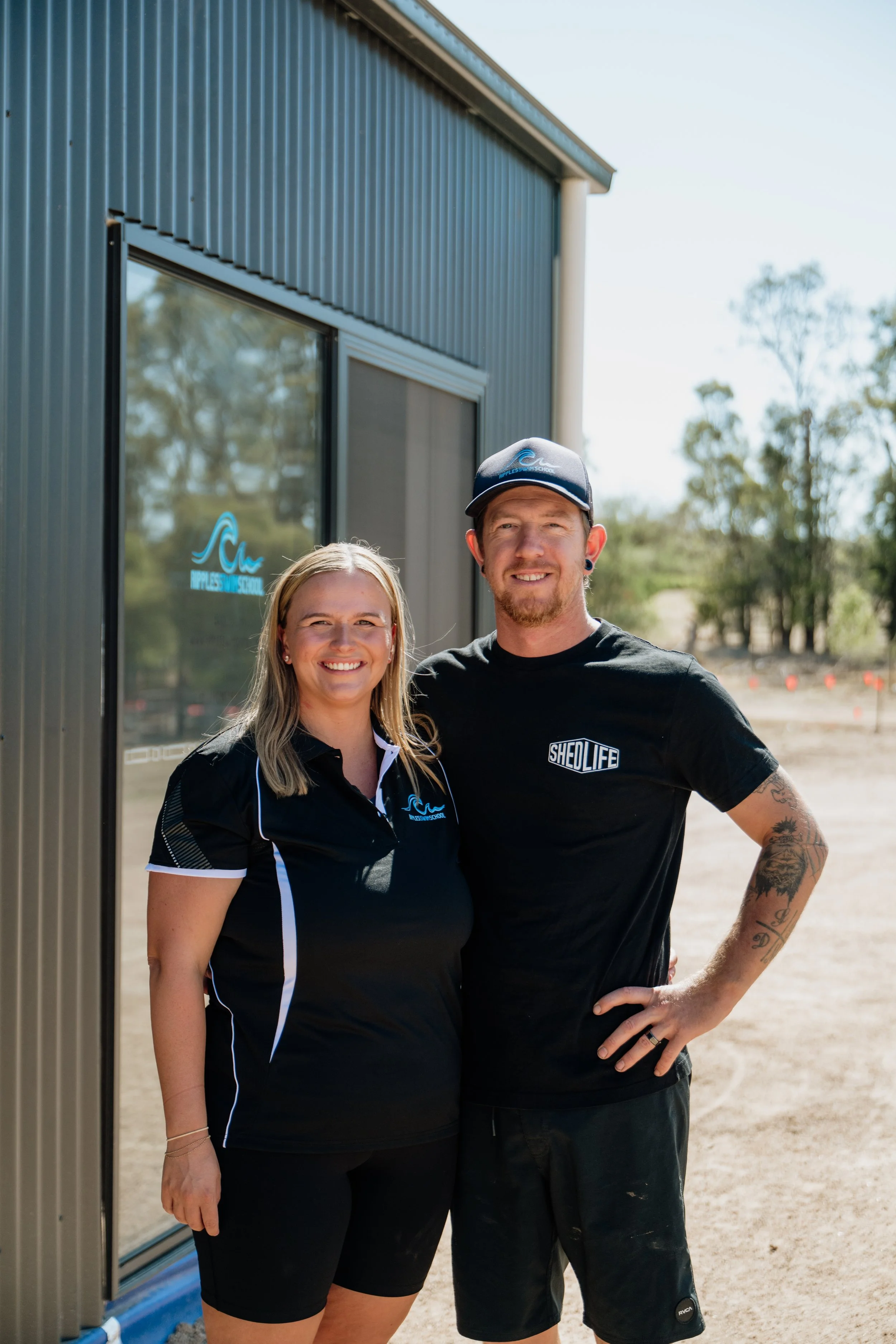 A man and woman standing outside a building with large glass windows, smiling at the camera. The woman is wearing a black polo shirt with a logo, and the man is wearing a black t-shirt with text and a cap. The background features a dirt area and trees.
