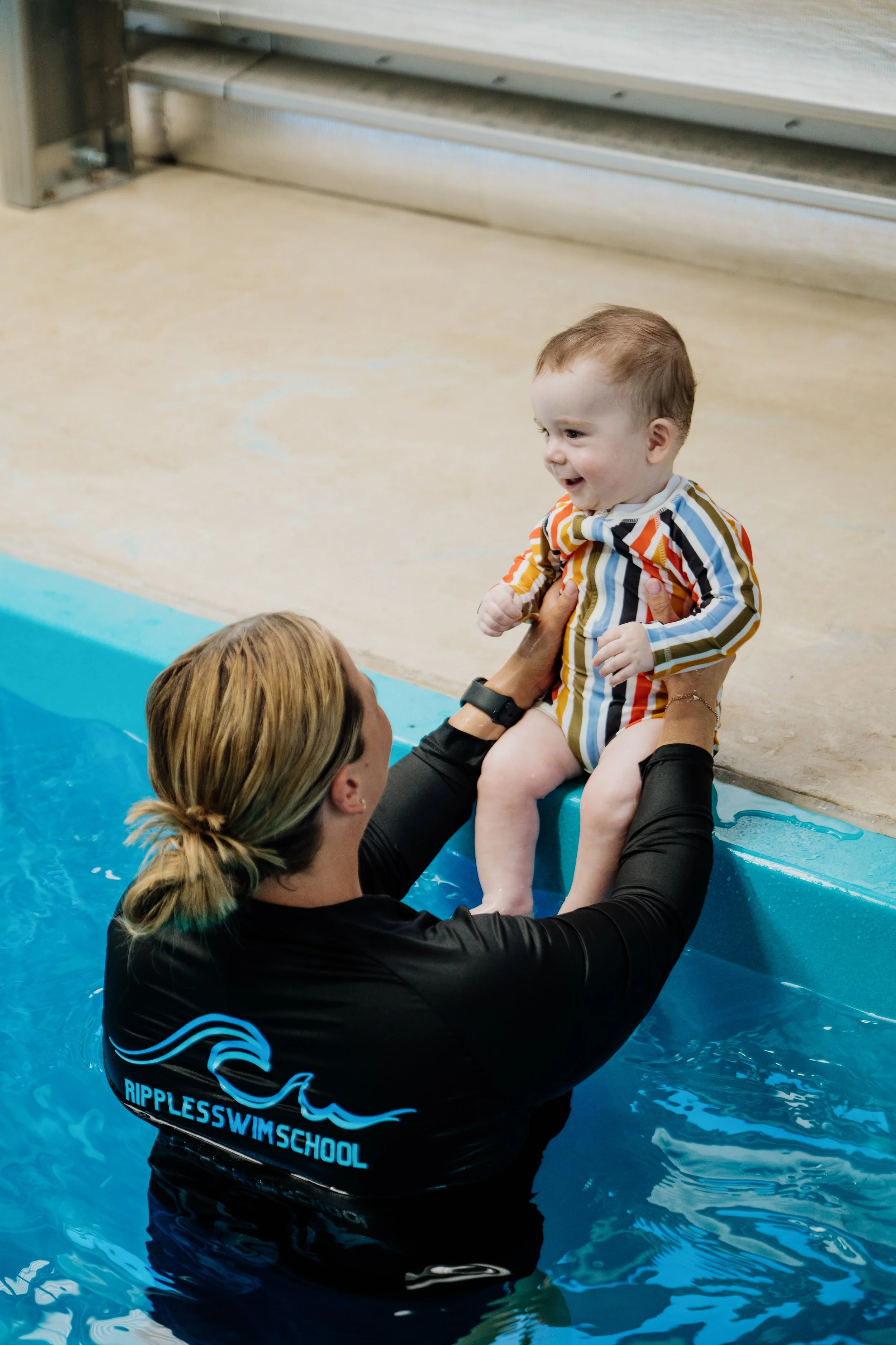 A woman in a black Ripples Swim School shirt holds a smiling baby with striped clothing at the edge of a swimming pool.