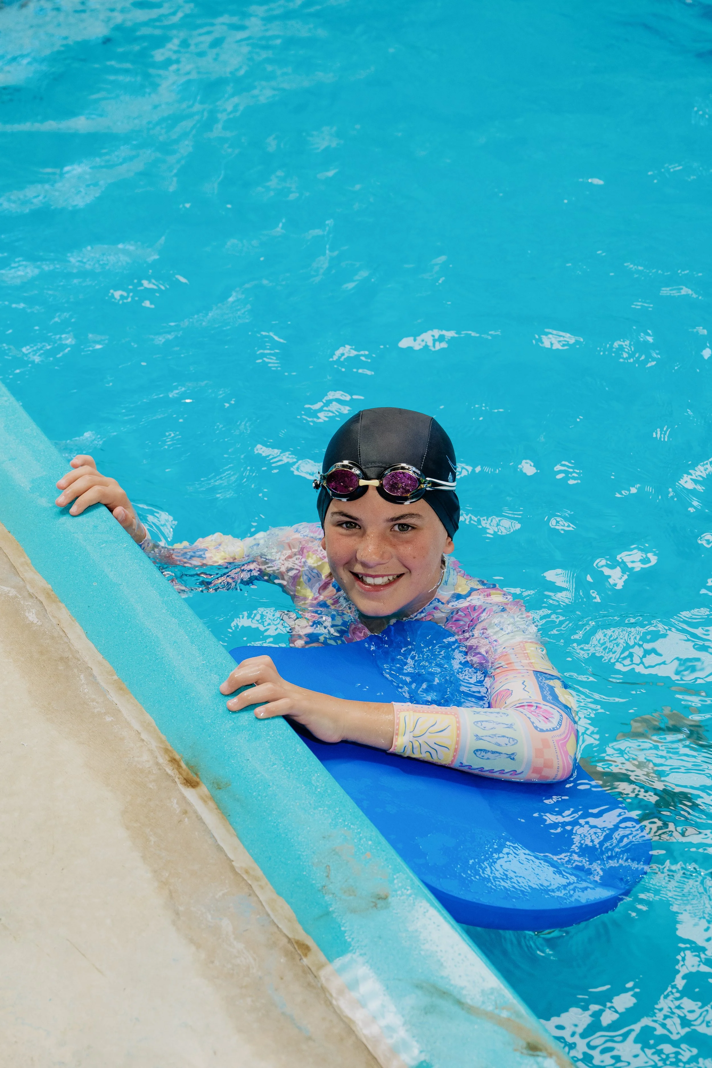 A young girl with goggles and a swim cap smiling in a swimming pool, holding a blue kickboard.