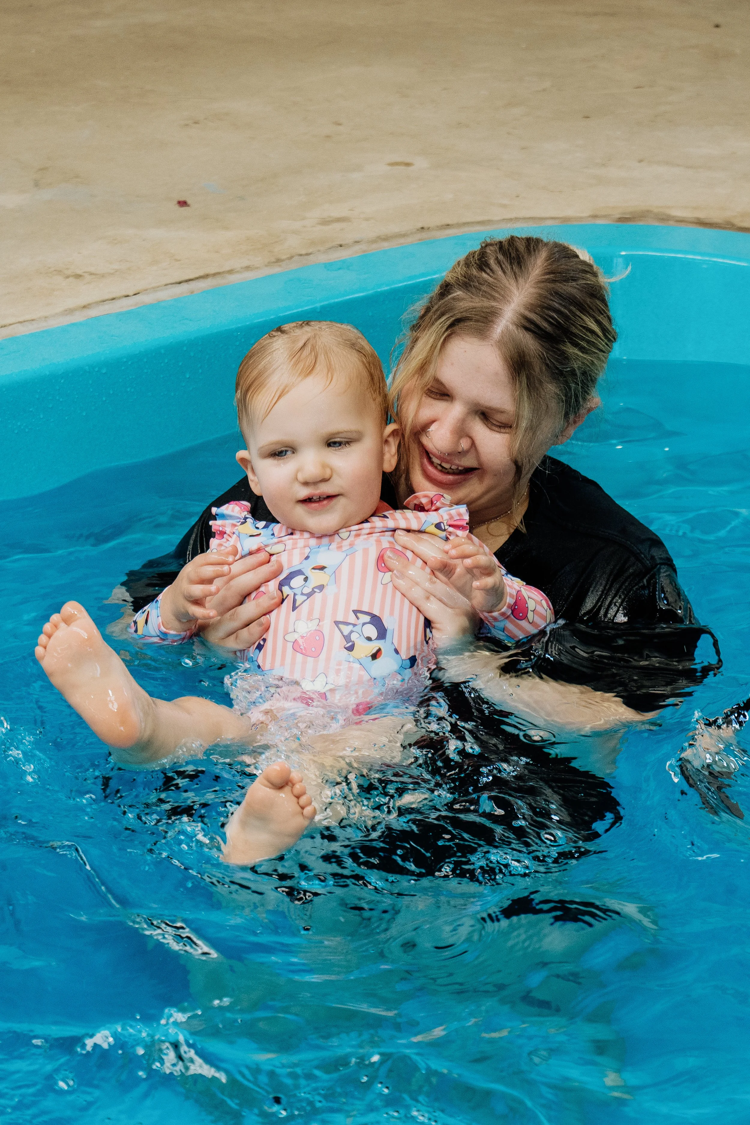 A woman is happily holding a young girl in a swimming pool, helping her float on her back. The girl wears a colorful swimsuit with cartoon characters. The scene is indoors with a concrete floor surrounding the pool.