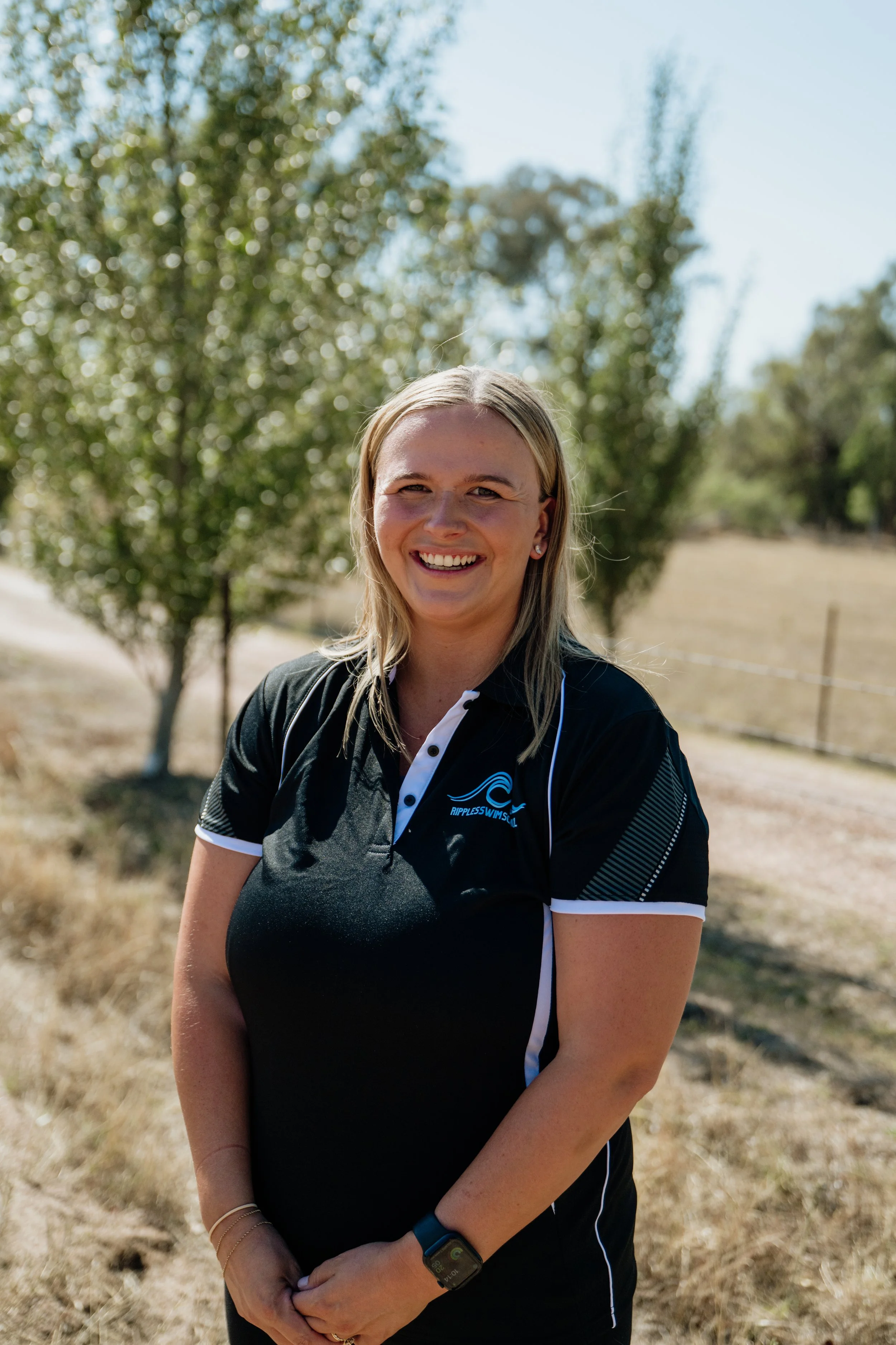 A woman is outdoors, smiling, wearing a black sports shirt with white accents and a logo for Ripples Swim School. She is standing on a grassy area with trees and a clear blue sky in the background.