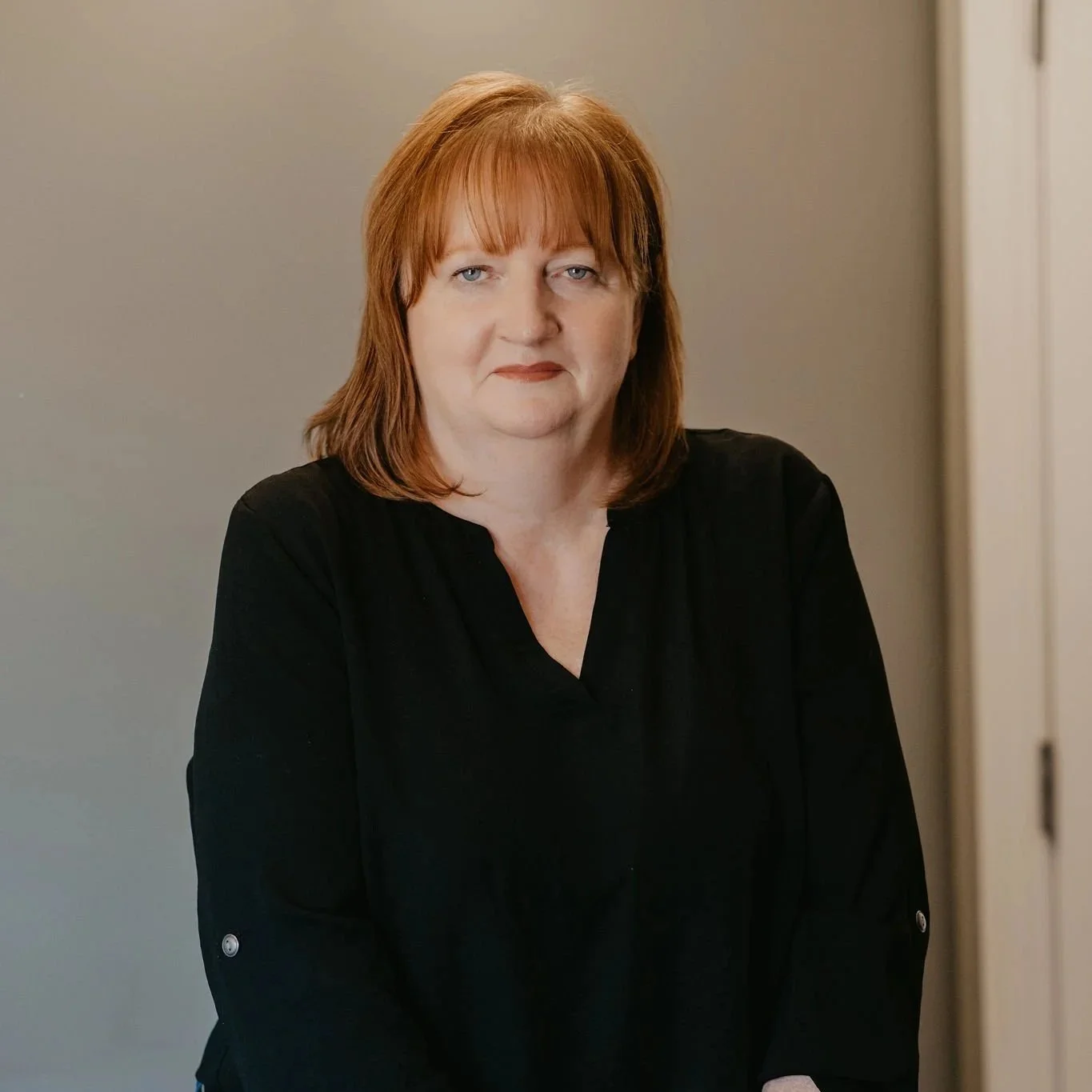 A middle-aged woman with reddish-brown hair and blue eyes, wearing a black top, standing indoors against a plain wall.