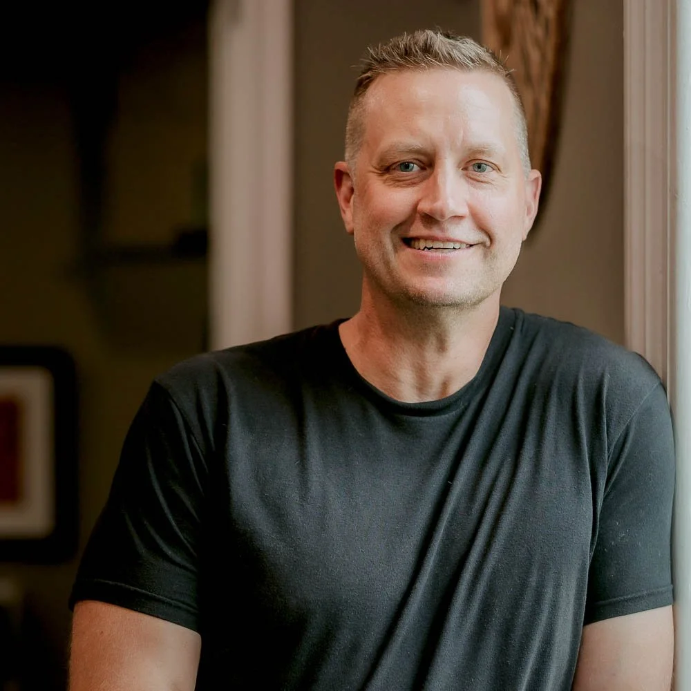 A smiling man with short blonde hair and a black t-shirt looking at the camera inside a house, near a window with curtains.