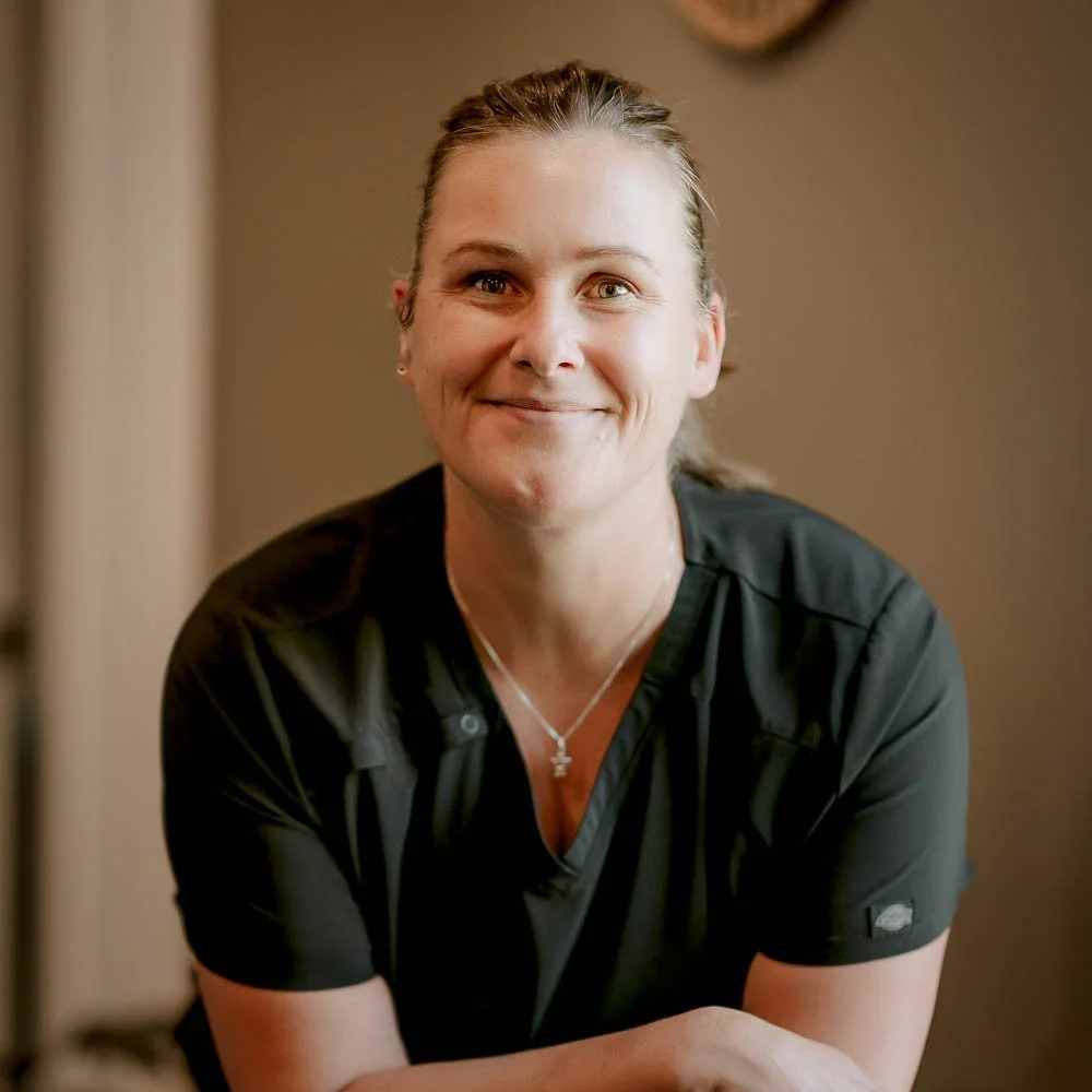A woman with light brown hair tied back, wearing a black shirt, smiling and looking at the camera against a beige wall background.
