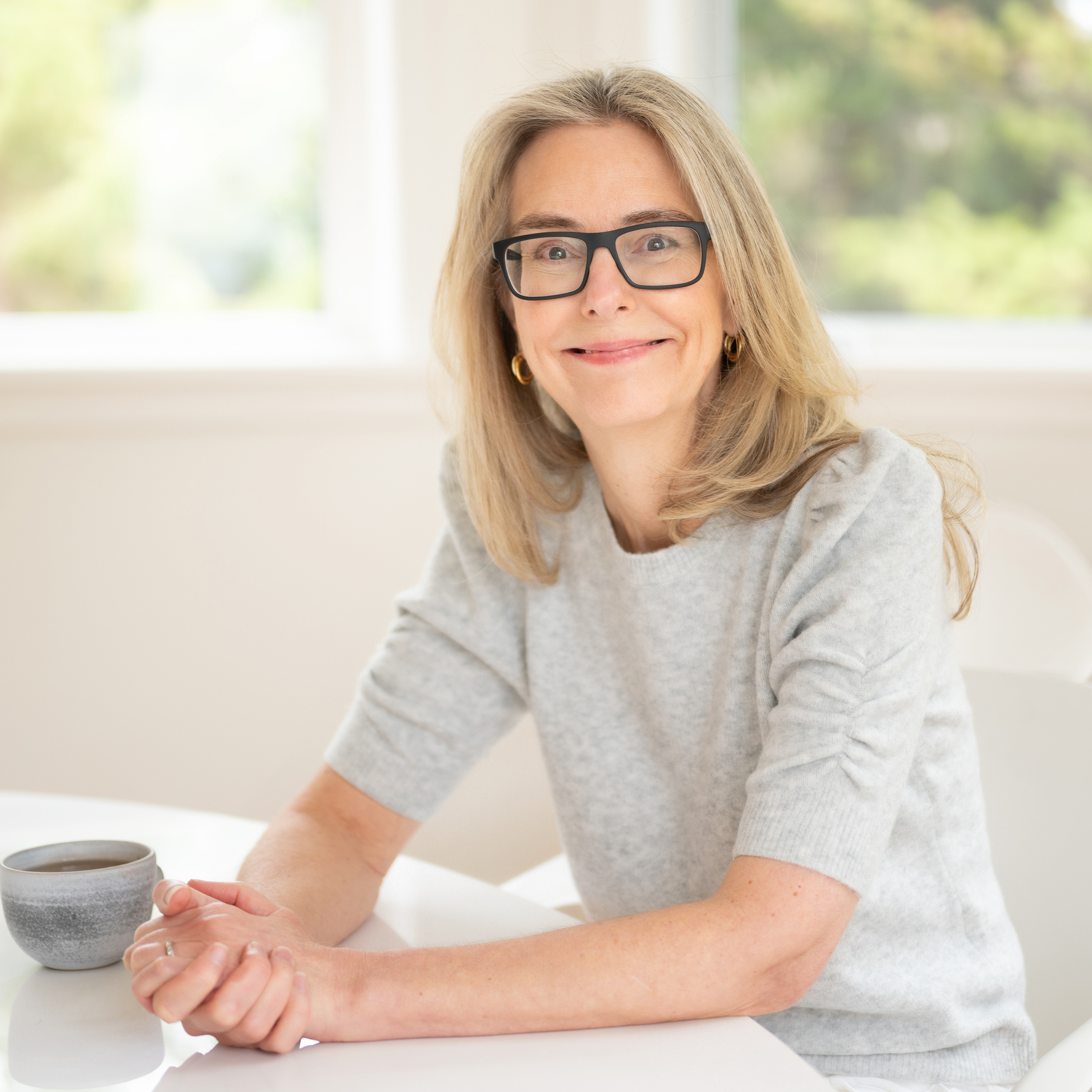 A woman with blonde hair, glasses, and a gray sweater is smiling while holding a gray mug of coffee or tea at a white table, with a window and greenery in the background.