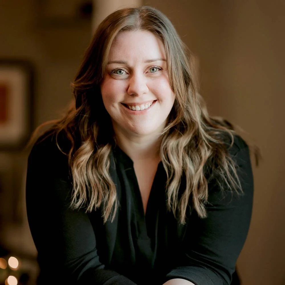 A woman with long, wavy brown hair and light-colored eyes smiling at the camera, wearing a black top in an indoor setting.