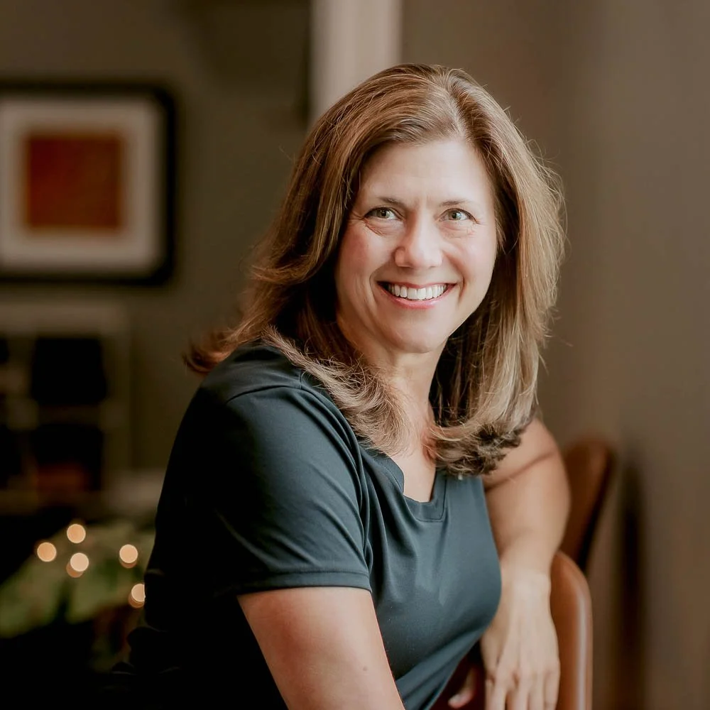A woman with light brown hair smiling, sitting indoors in a black shirt, with blurred framed art and lights in the background.
