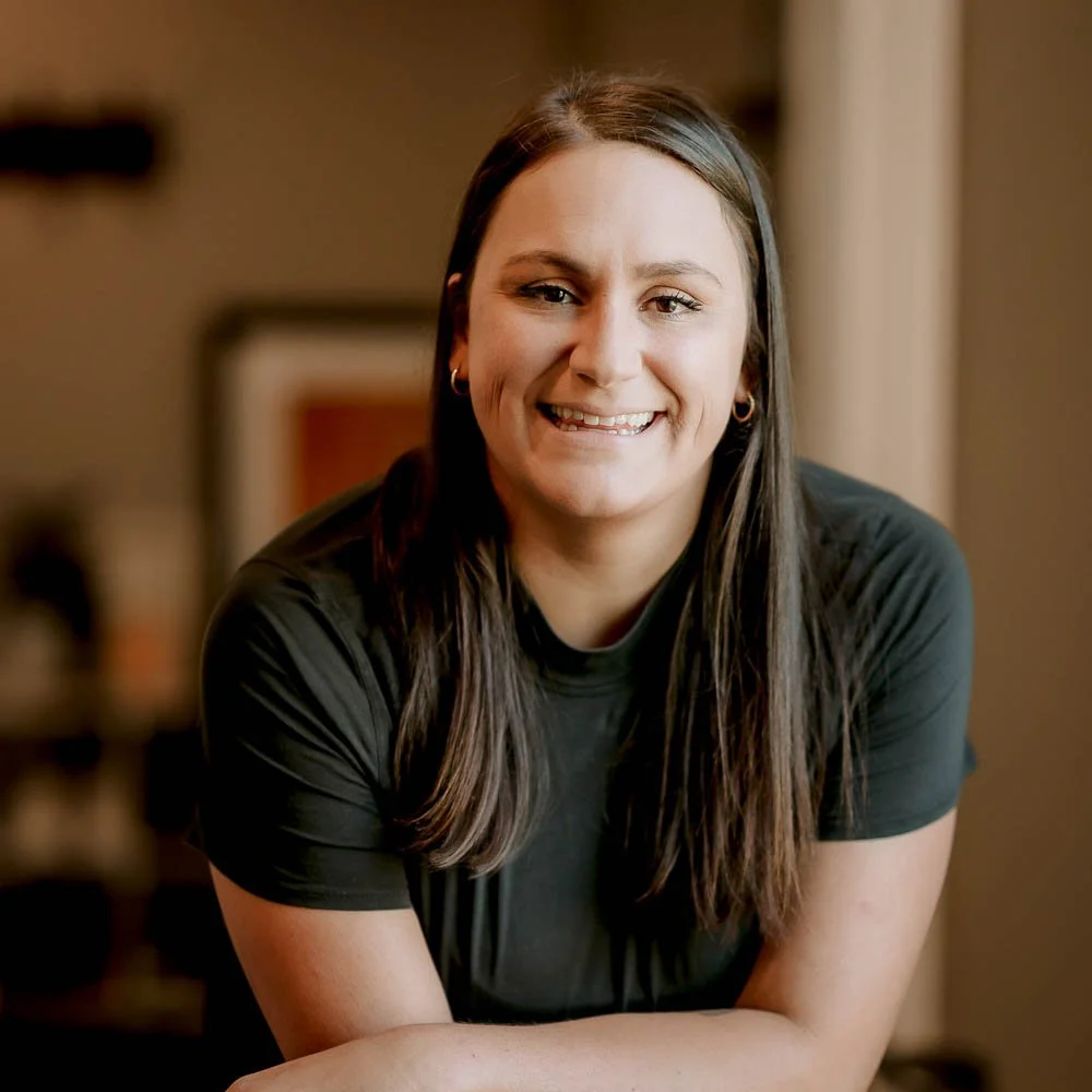 A smiling young woman with long dark hair and hoop earrings, wearing a black t-shirt, sitting indoors with a blurred background.