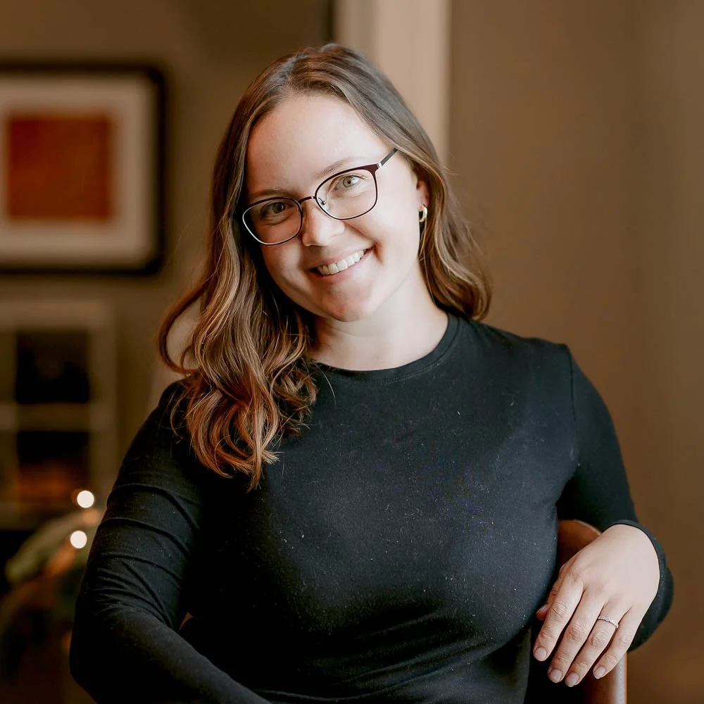 Woman with glasses smiling, wearing a black long-sleeve shirt, sitting indoors with a blurred background.