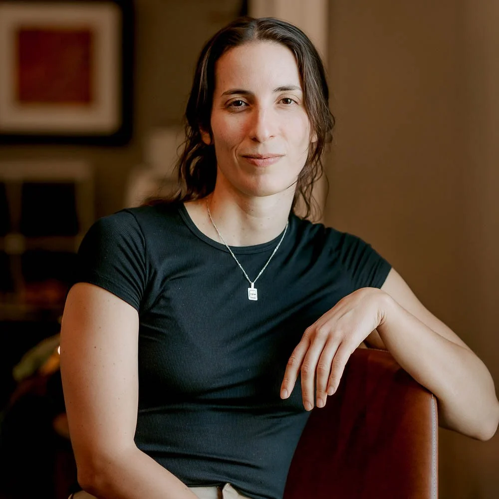 A woman with shoulder-length brown hair, wearing a black t-shirt and a silver necklace, sitting indoors with her right arm resting on a piece of furniture, smiling softly at the camera.