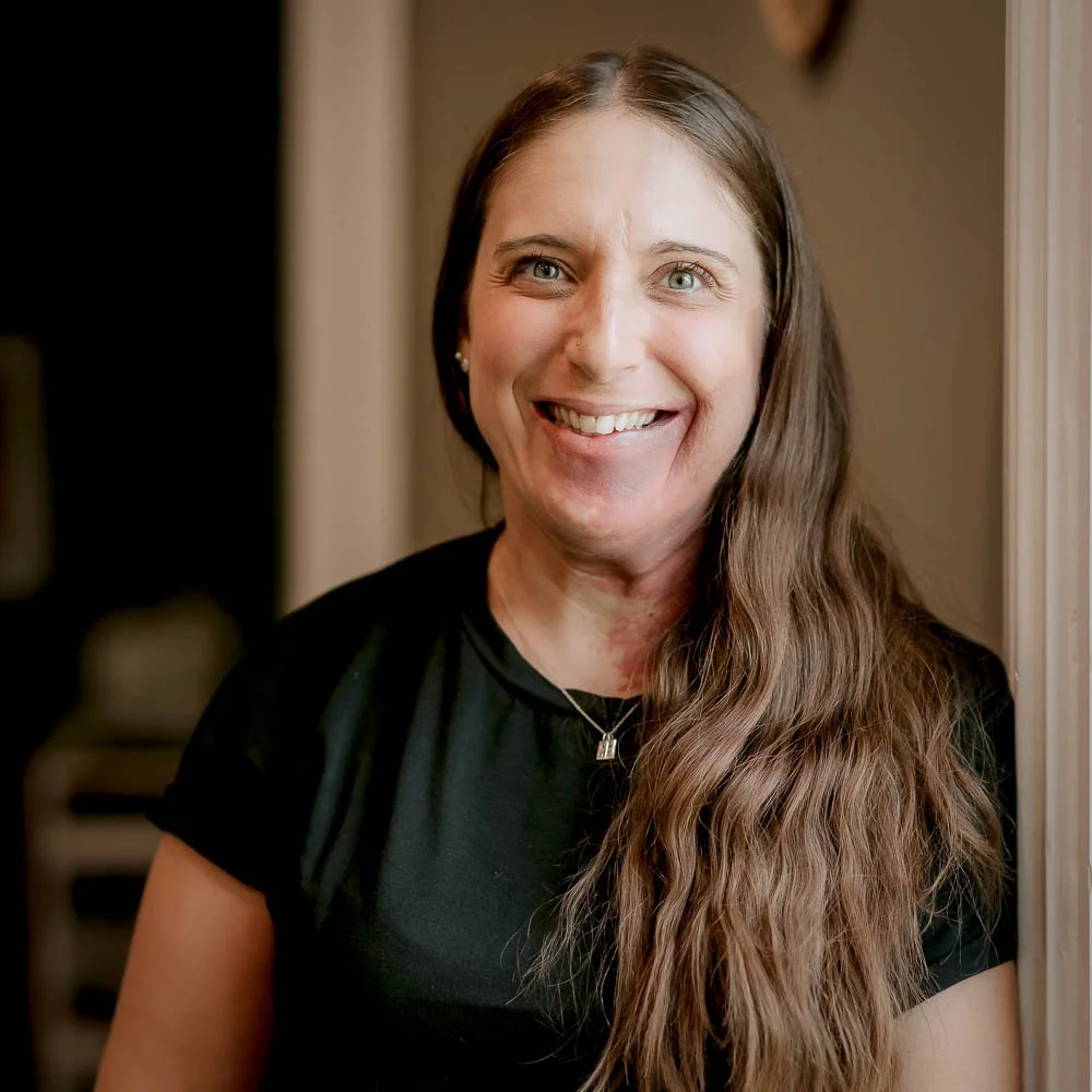 A woman with long brown hair, blue eyes, and a big smile posing indoors near a wall, wearing a black shirt and small earrings.