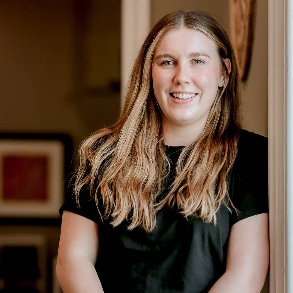 A young woman with long, wavy blonde hair, wearing a black t-shirt, smiling and standing indoors near a doorway
