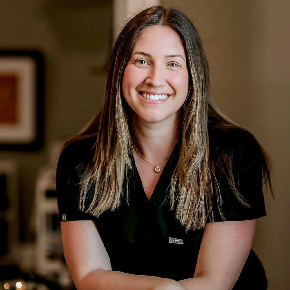 A smiling woman with long brown hair, wearing a black shirt and a gold necklace, posing indoors.